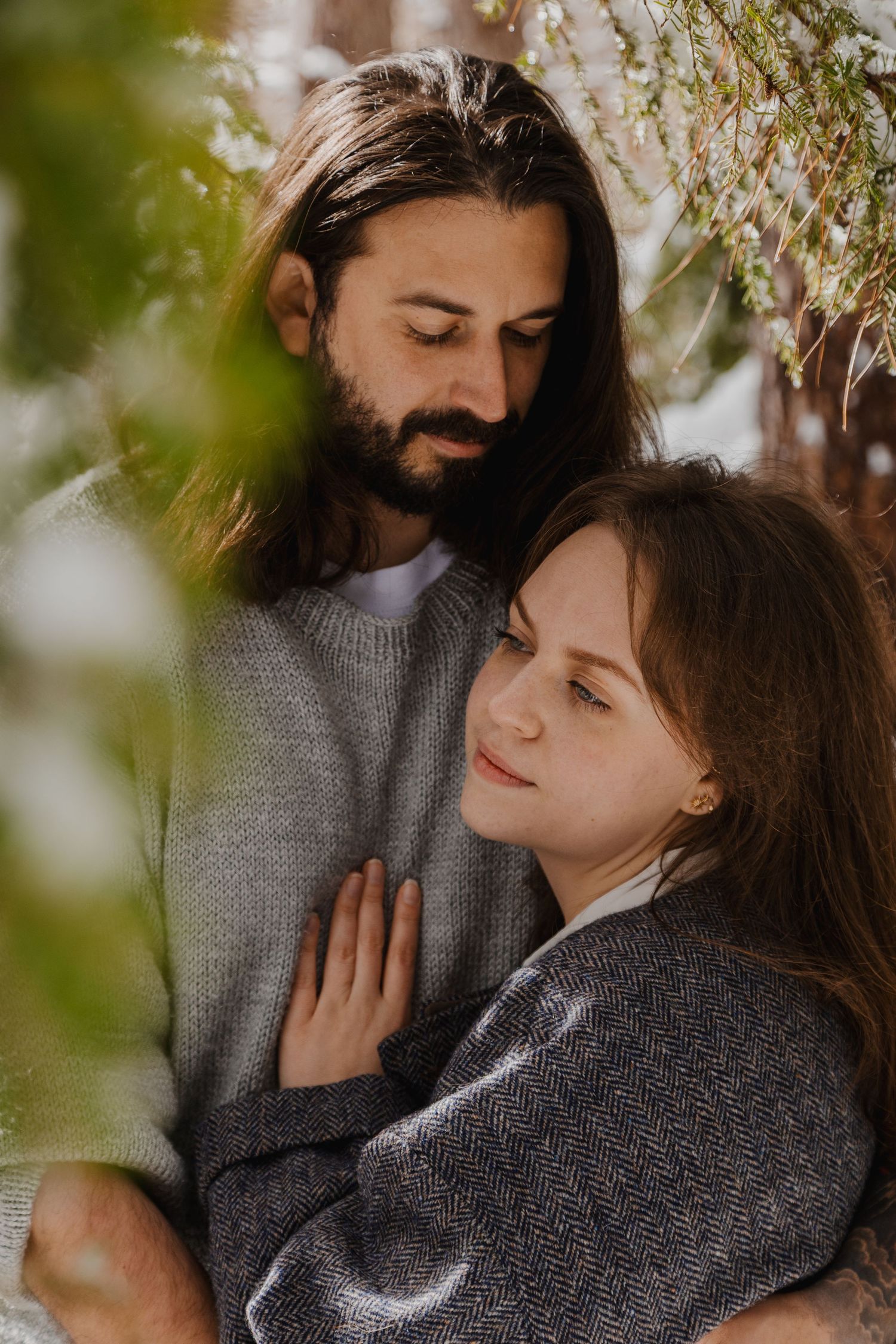 A bearded man with long hair embraces a young woman in gray sweaters outdoors among blooming branches.