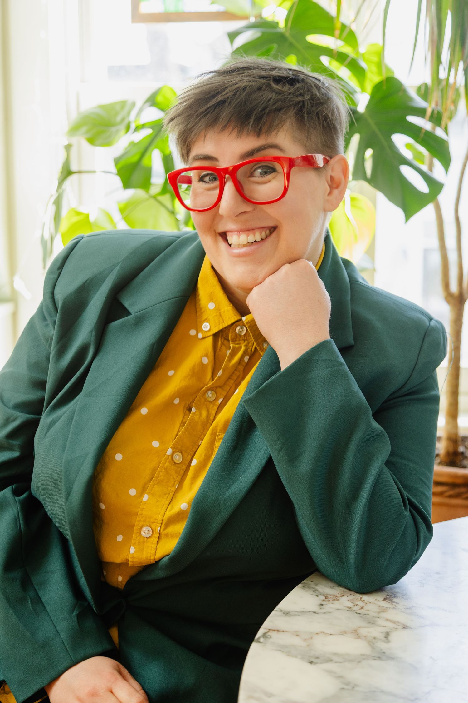 Casual portrait with red glasses, green blazer and yellow polka dot shirt near indoor plants.