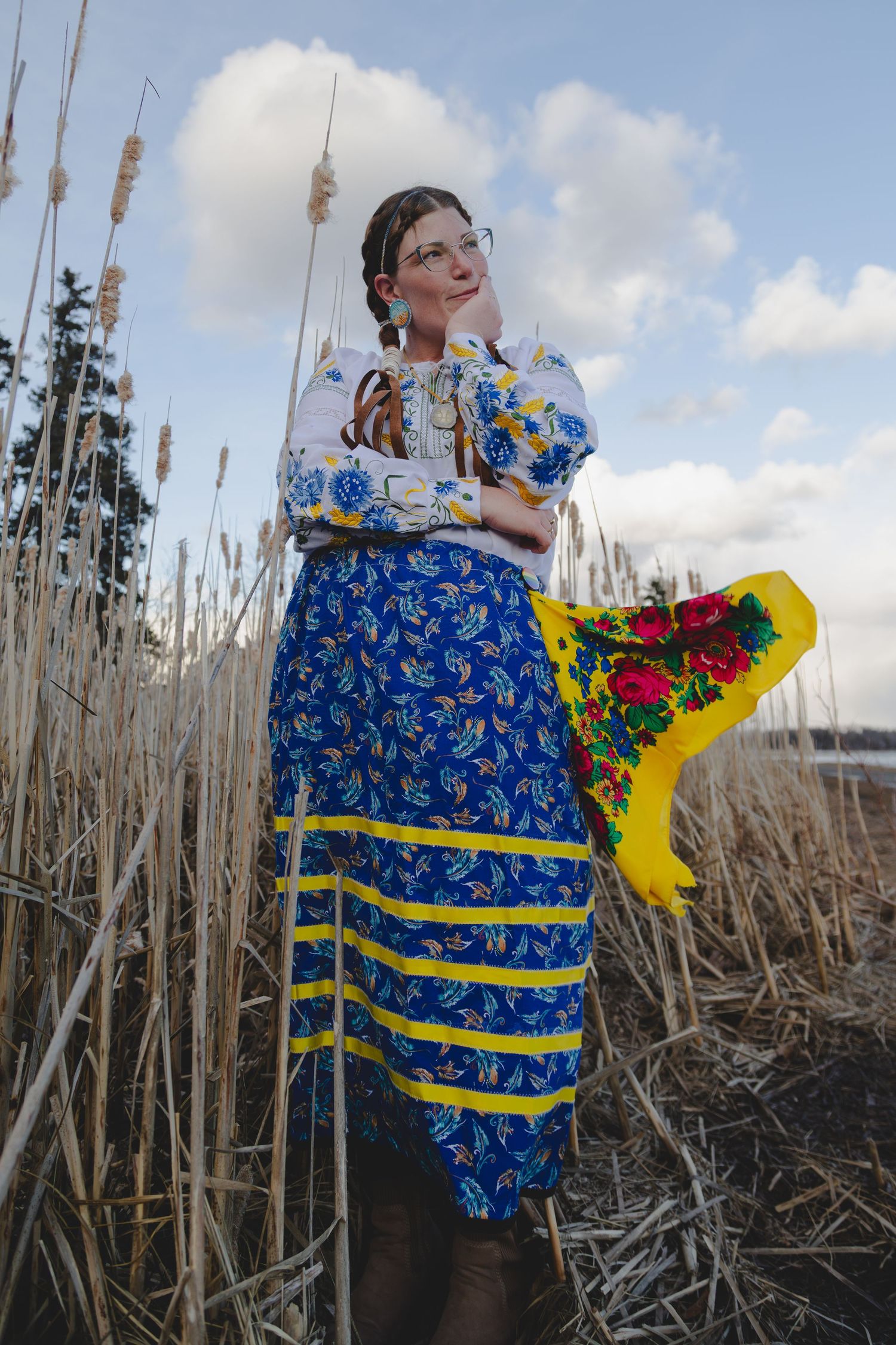 Woman in traditional Ukrainian embroidered blouse and blue floral skirt holding colorful bouquet among reeds.