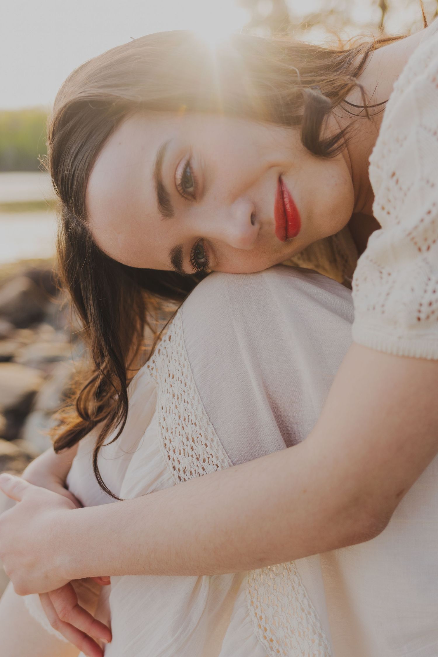 Young woman with red lips in white lace dress sitting outdoors, golden backlit sunset behind her.