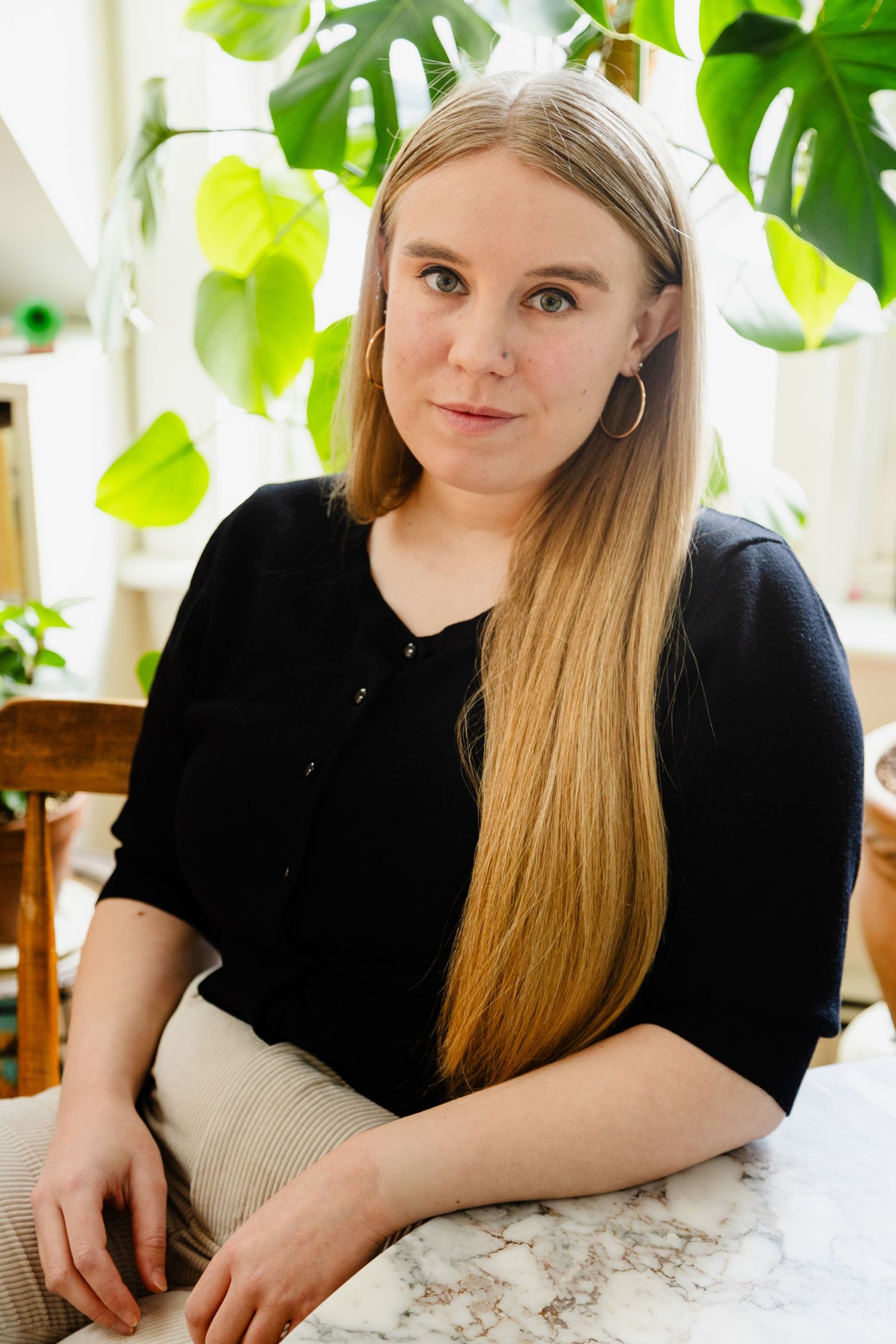 A person in a black top sits at a marble table with lush green monstera plants in the background.