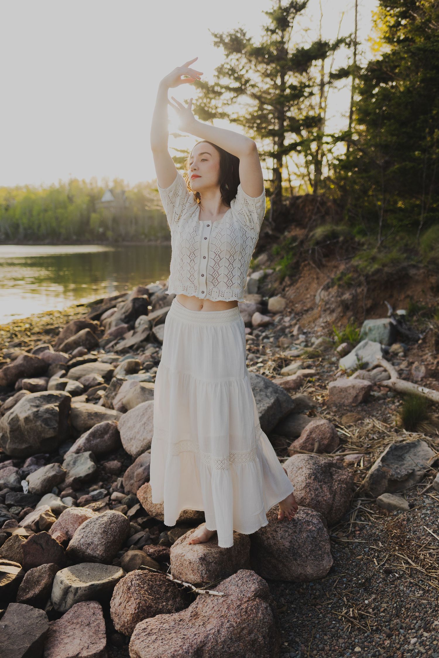 Woman in white crop top and maxi skirt poses gracefully on rocky lakeshore at golden hour sunset.