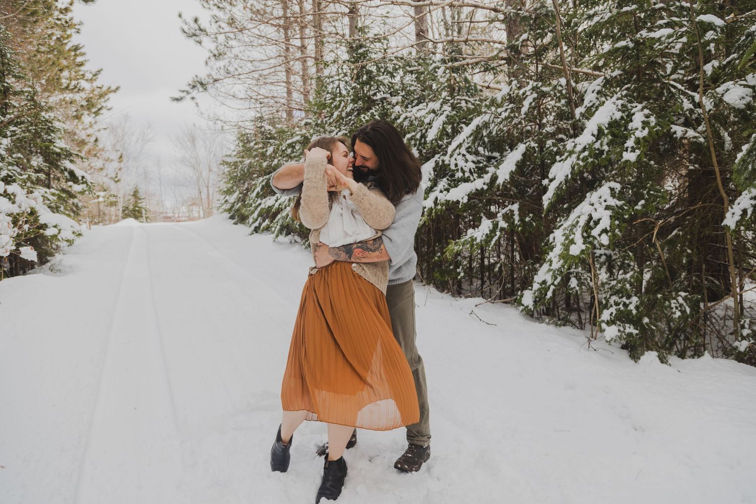 Couple embracing and dancing on a snowy forest road in winter, surrounded by snow-covered evergreen trees.