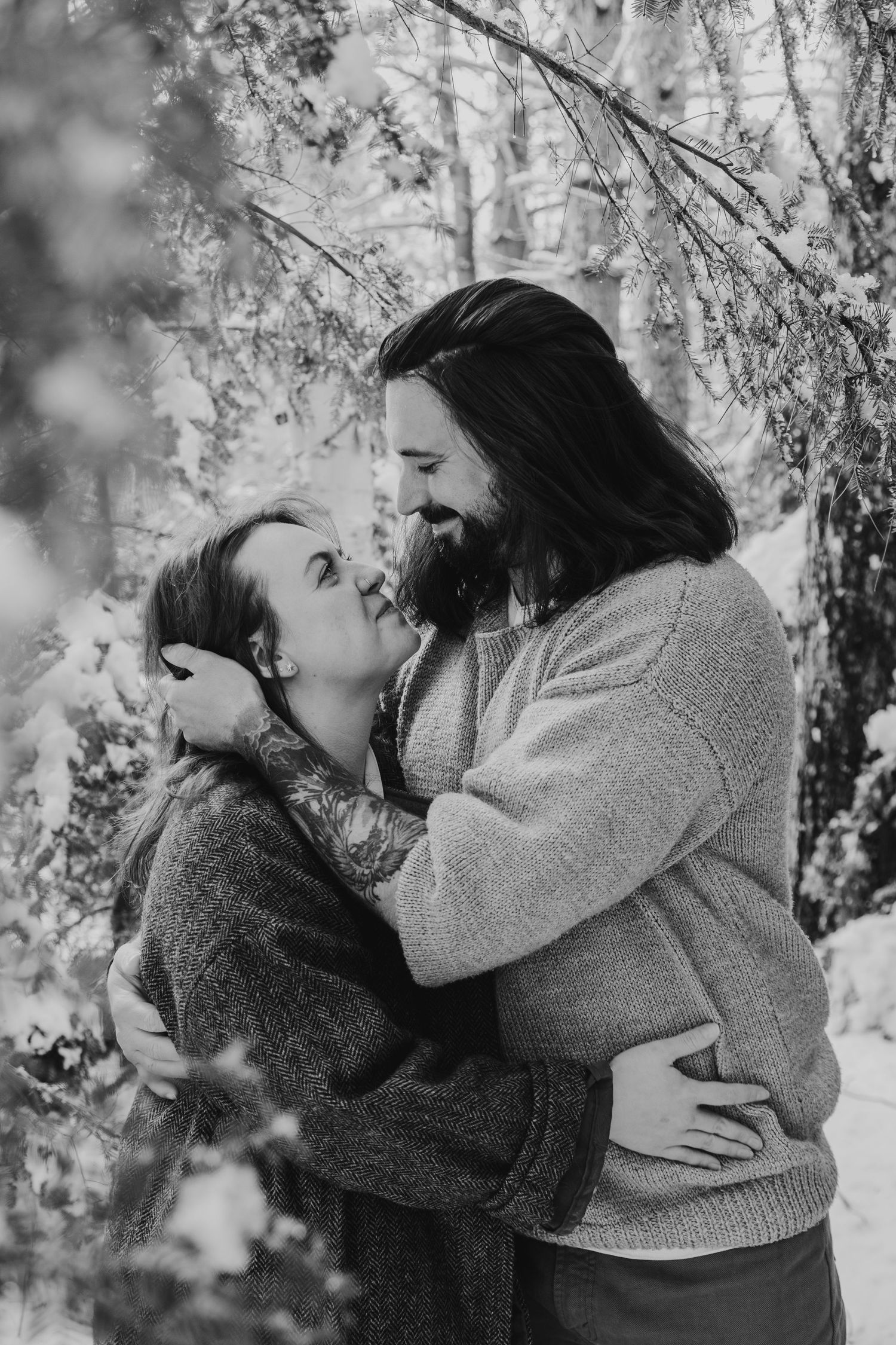 Black and white photo of a couple embracing and about to kiss in a snowy winter forest setting.