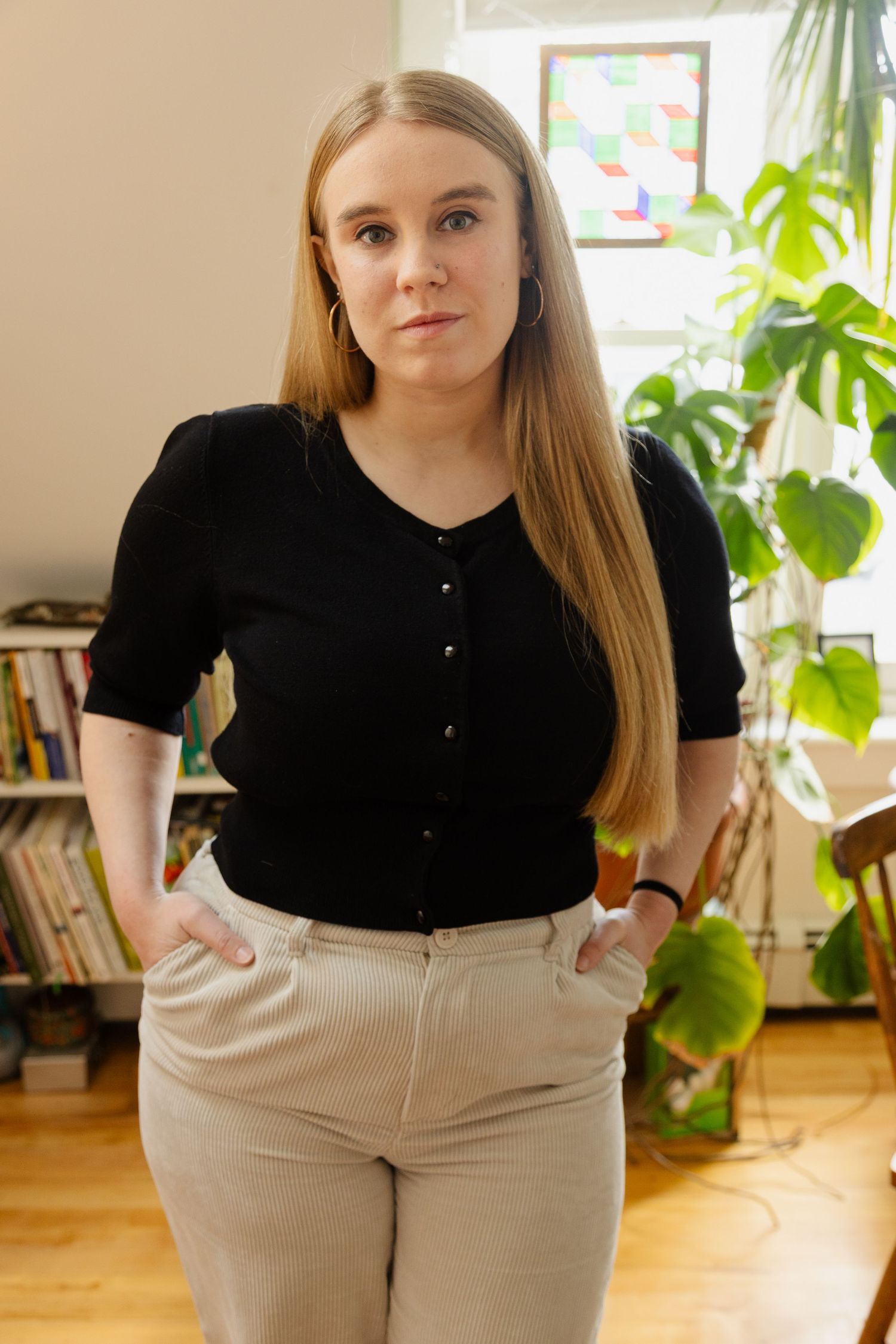 A person wearing a black cardigan and beige pants stands in a room with houseplants and bookshelves.