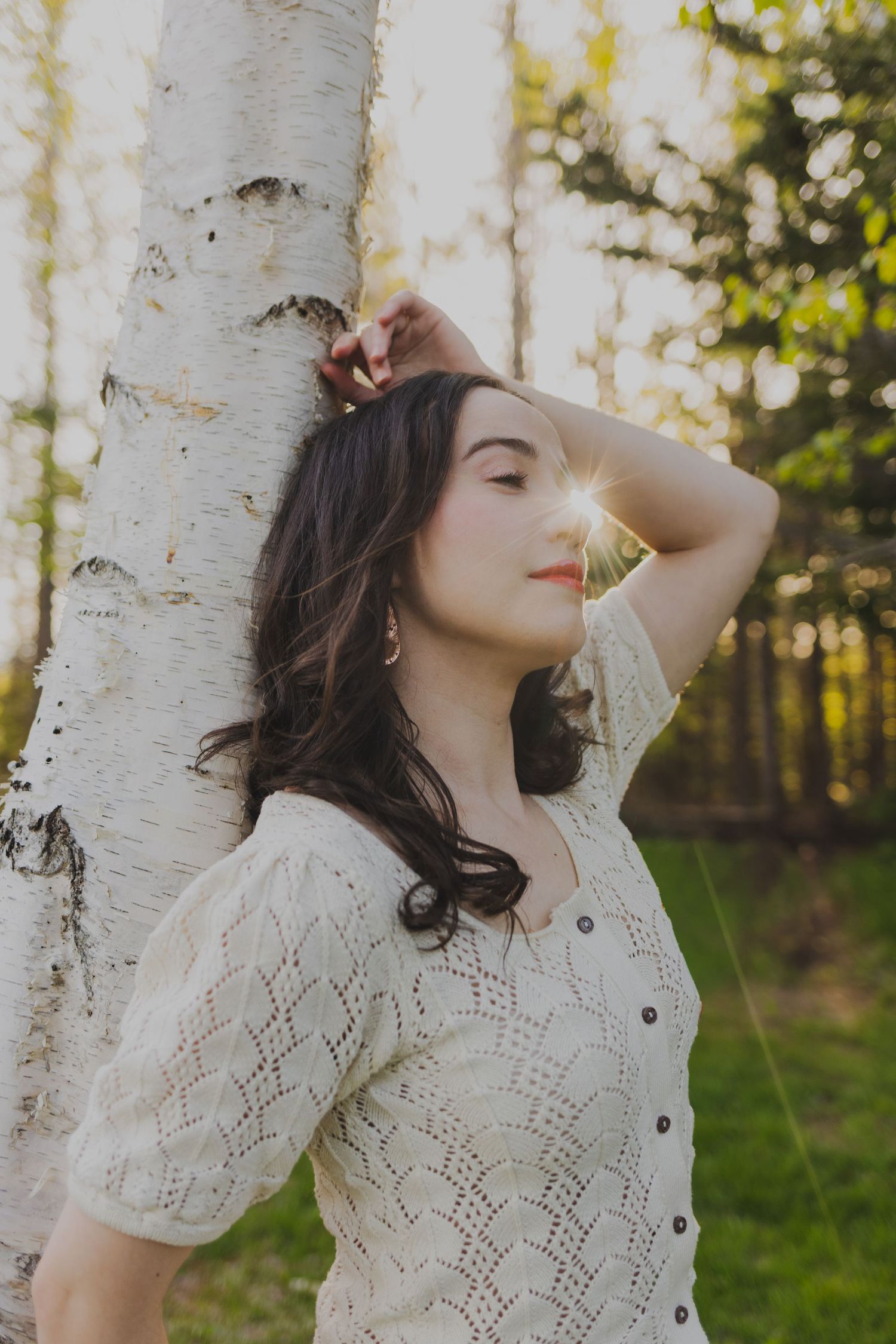 Young woman in white lace top leaning against birch tree with golden sunlight in forest setting.