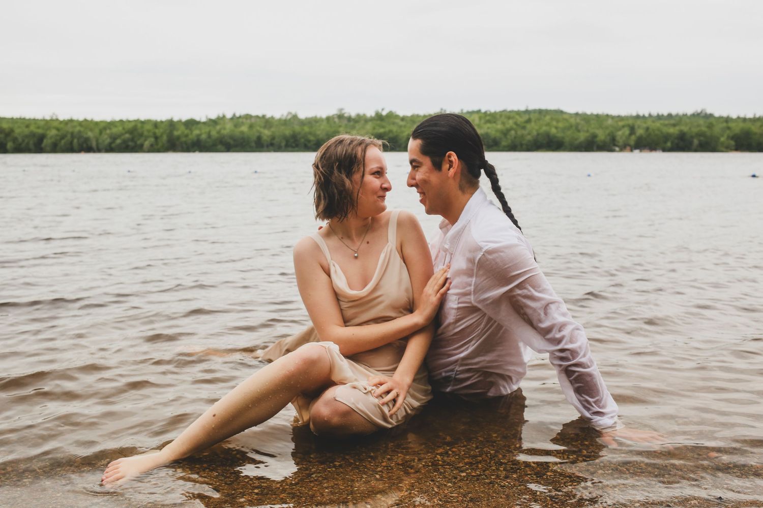 Couple sitting together in shallow lake water, sharing an intimate moment with lush green trees in the background.