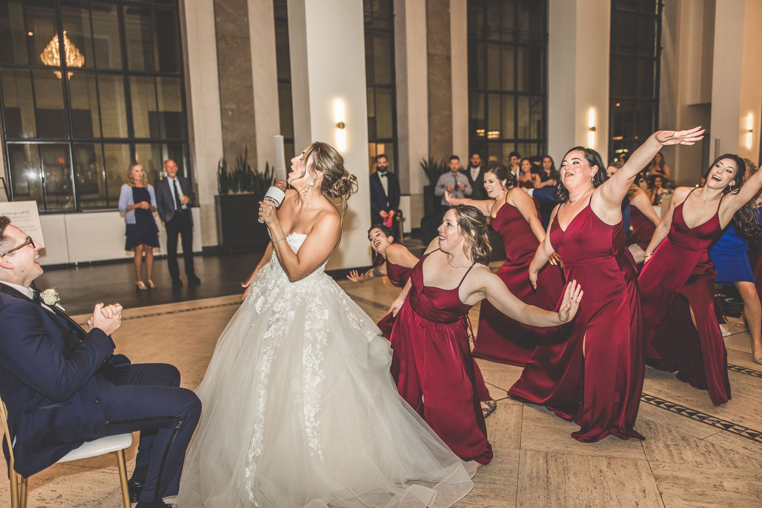 Wedding party performs choreographed dance routine with bride in white gown and bridesmaids in burgundy dresses.