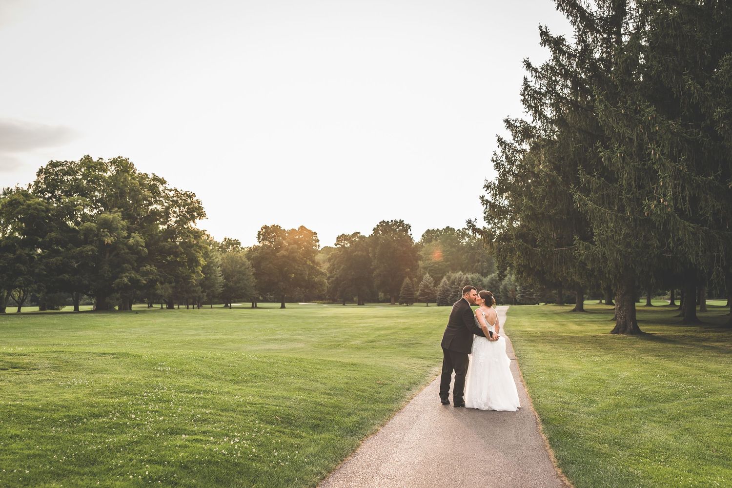 Wedding couple silhouetted against sunset on tree-lined golf course path.