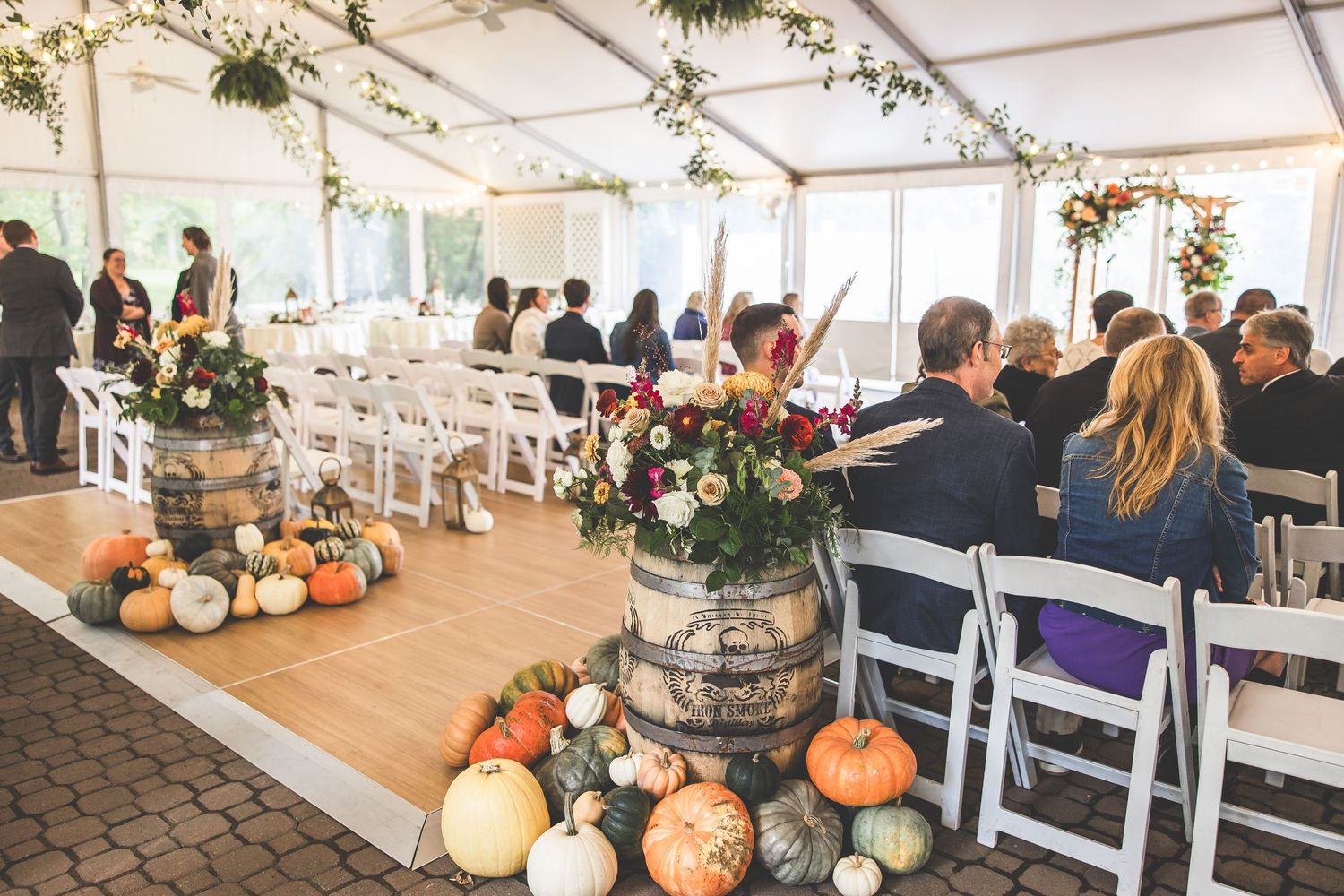 Wedding ceremony setup under tent with white chairs and rustic barrel flower arrangements with pumpkins.