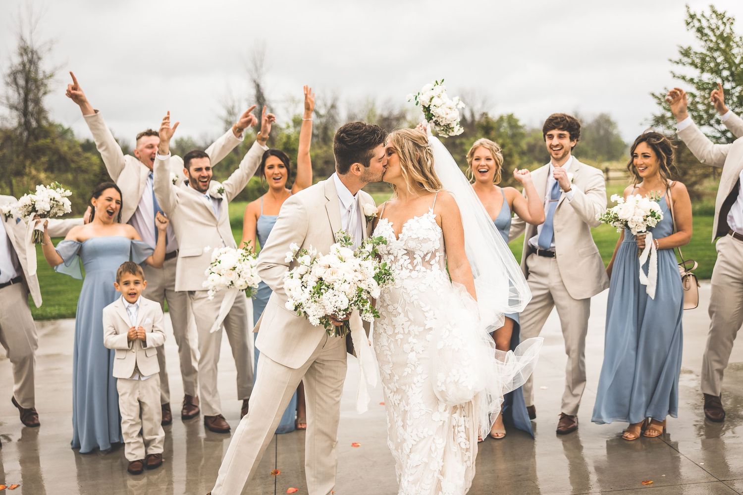 Wedding party celebrates with raised arms while couple walks through on rainy outdoor patio.