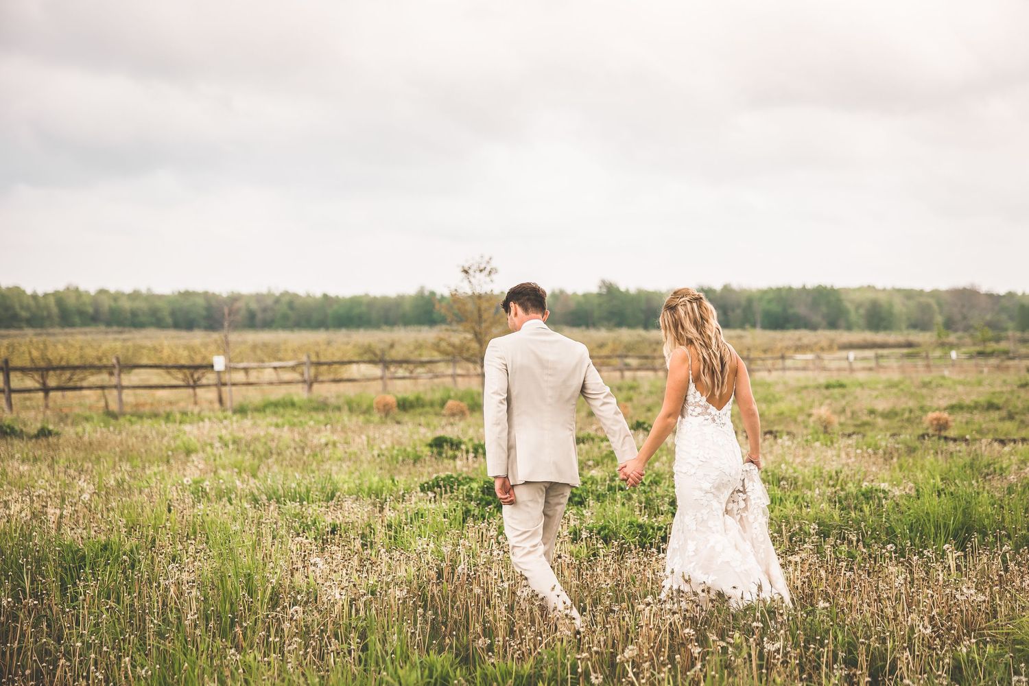 A romantic couple walks through a sunlit meadow of wildflowers wearing white and beige wedding attire.