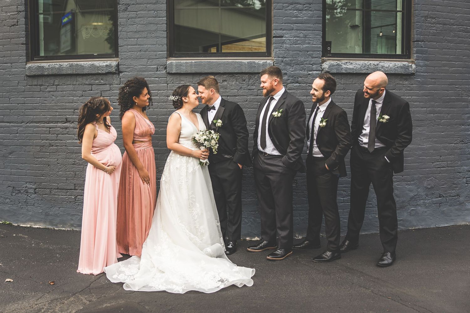 Wedding party in pink and black attire posing against industrial grey brick wall.