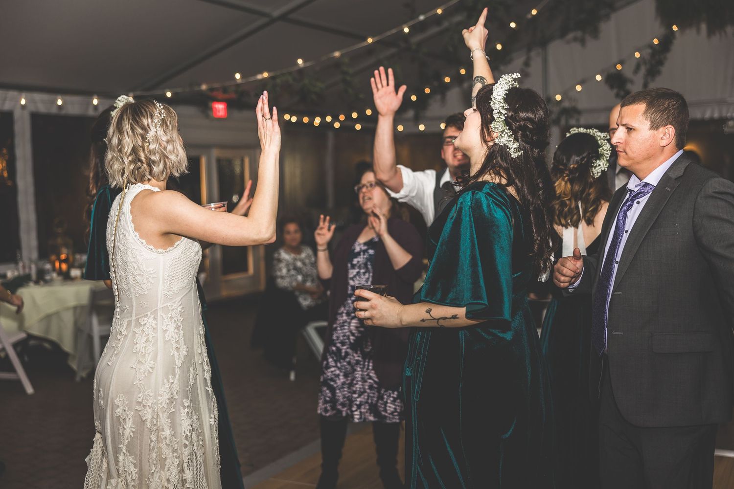 Wedding reception guests dancing and celebrating under decorative string lights in an indoor venue.