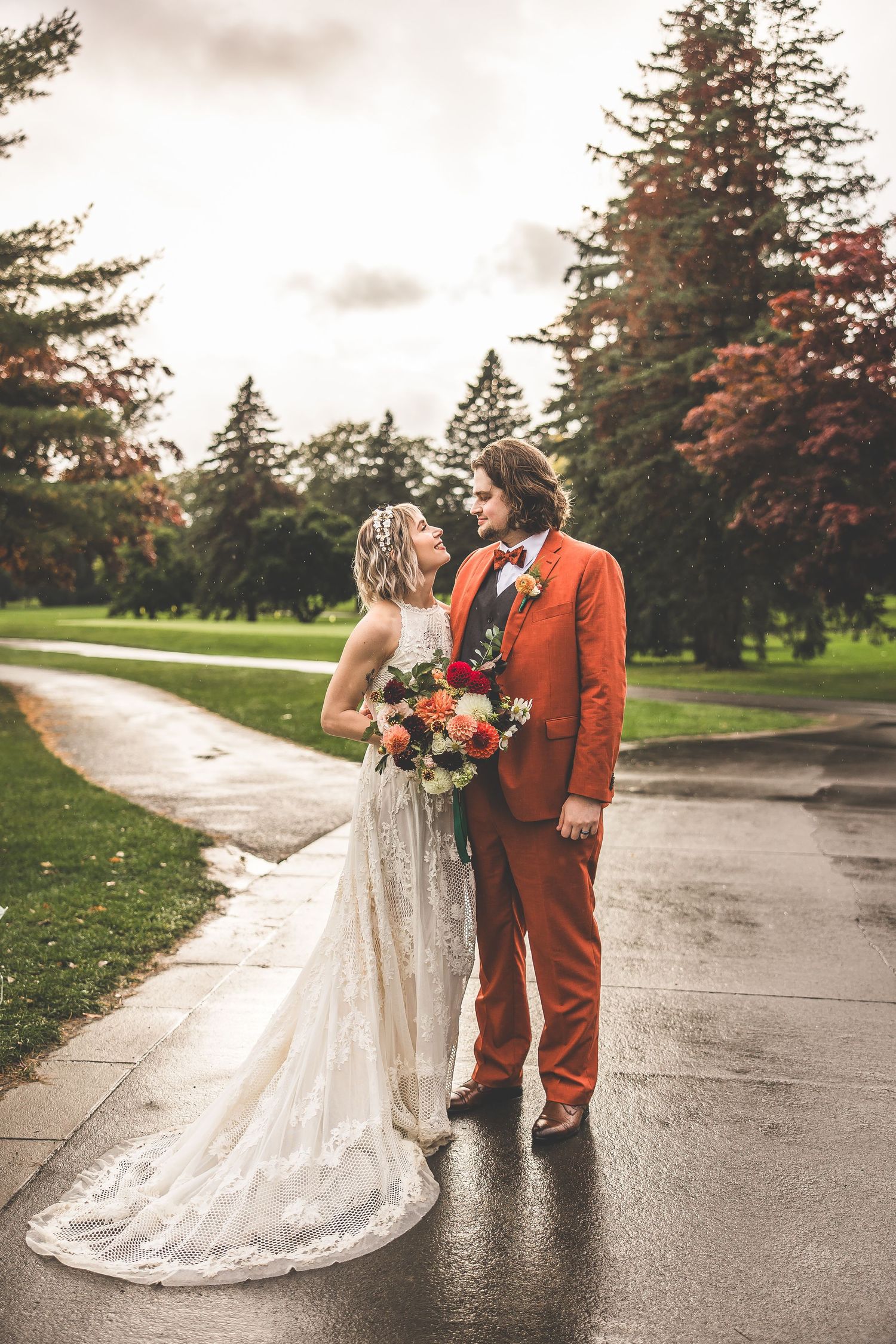 Wedding couple in white dress and orange suit walking on wet pavement surrounded by evergreen trees.