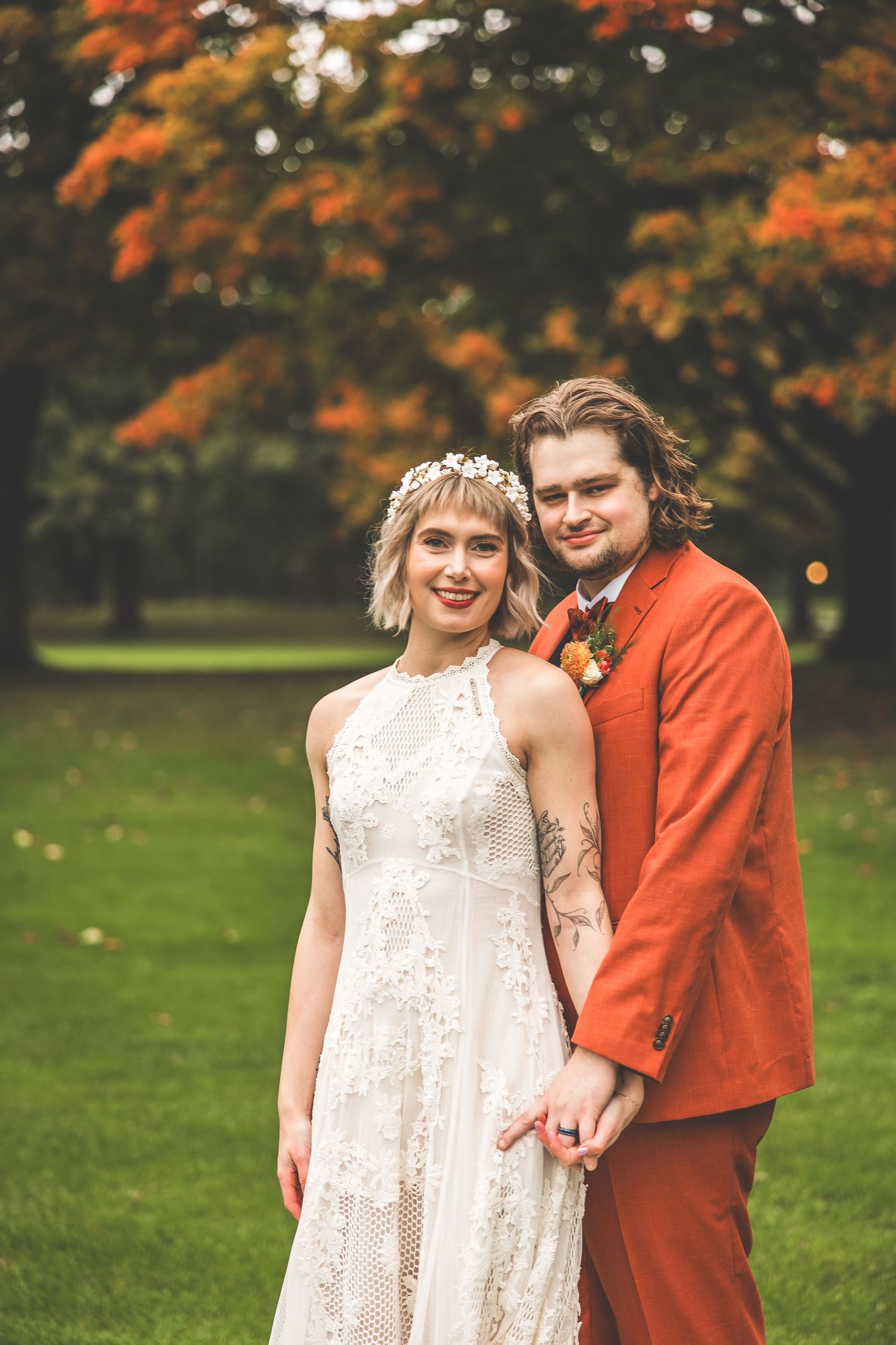 A couple poses in a garden with autumn foliage wearing a white lace dress and burnt orange suit.