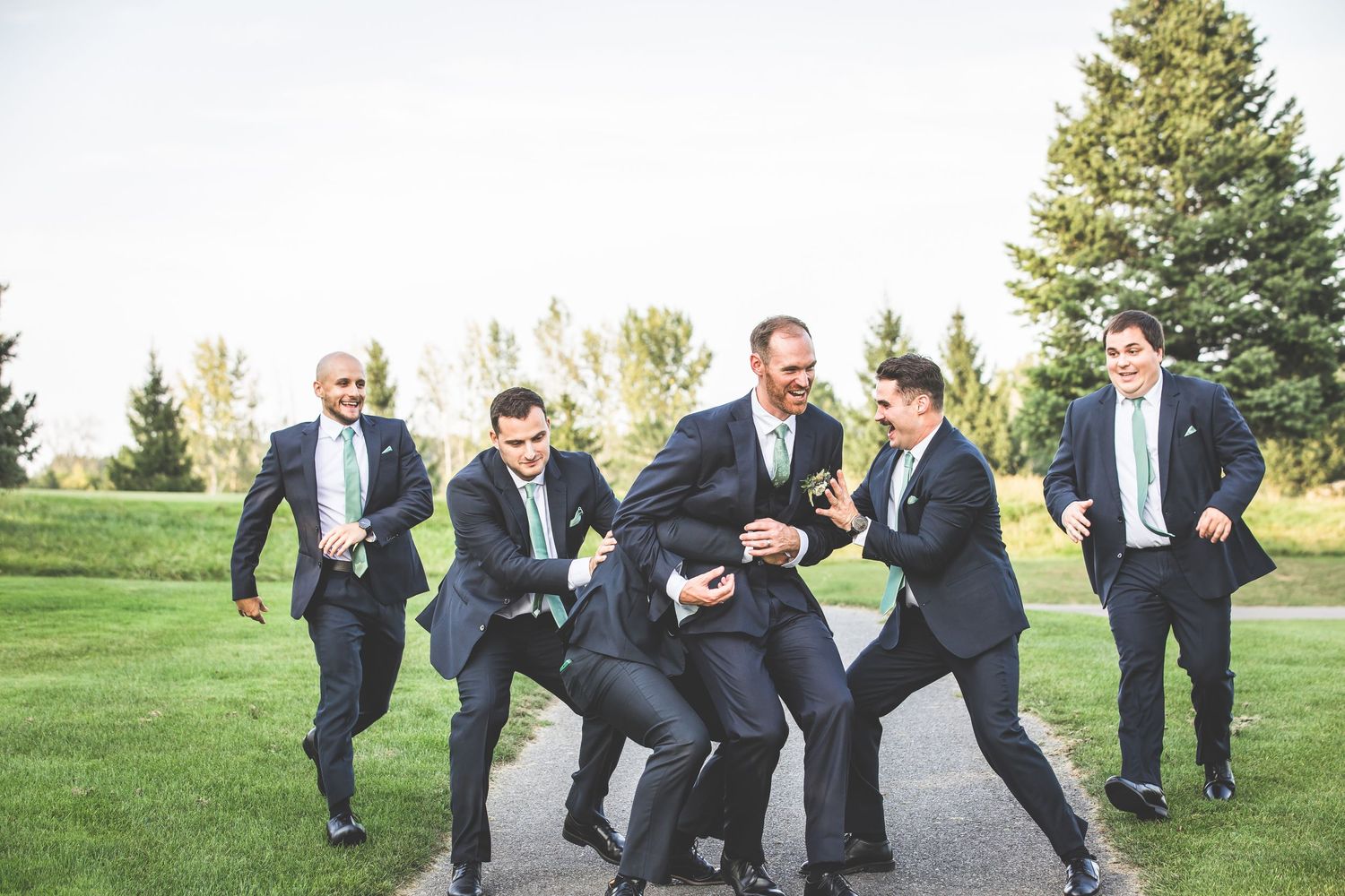 Groomsmen playfully roughhouse on grass during wedding photo session.