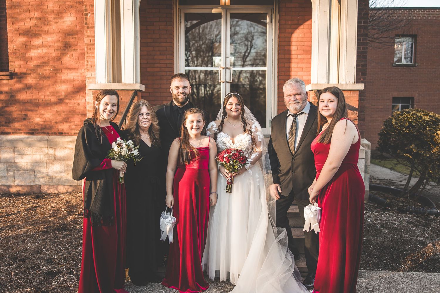 Wedding party in red and black attire gathered outside historic brick church.