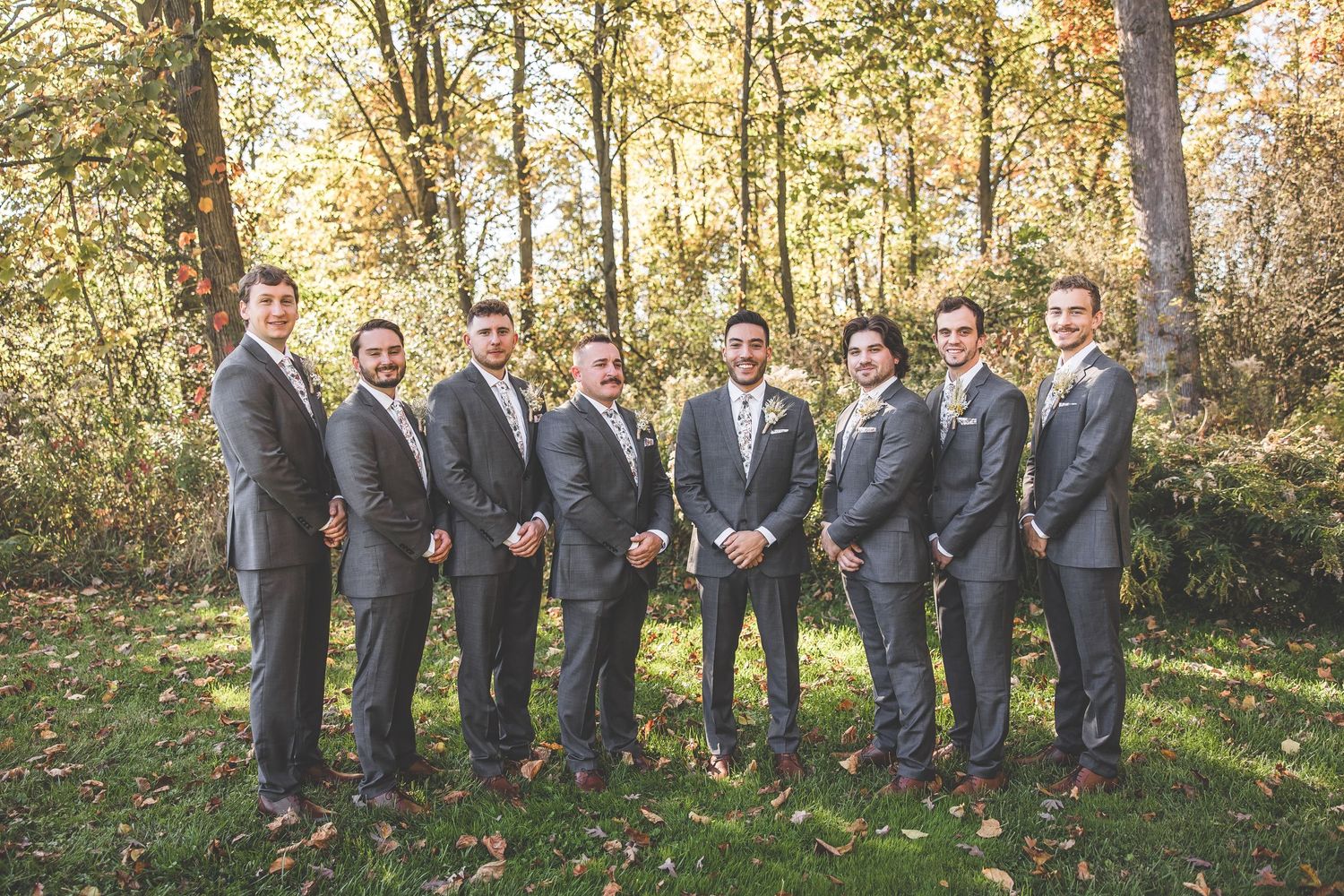Groomsmen in matching gray suits pose together in an autumn forest setting.