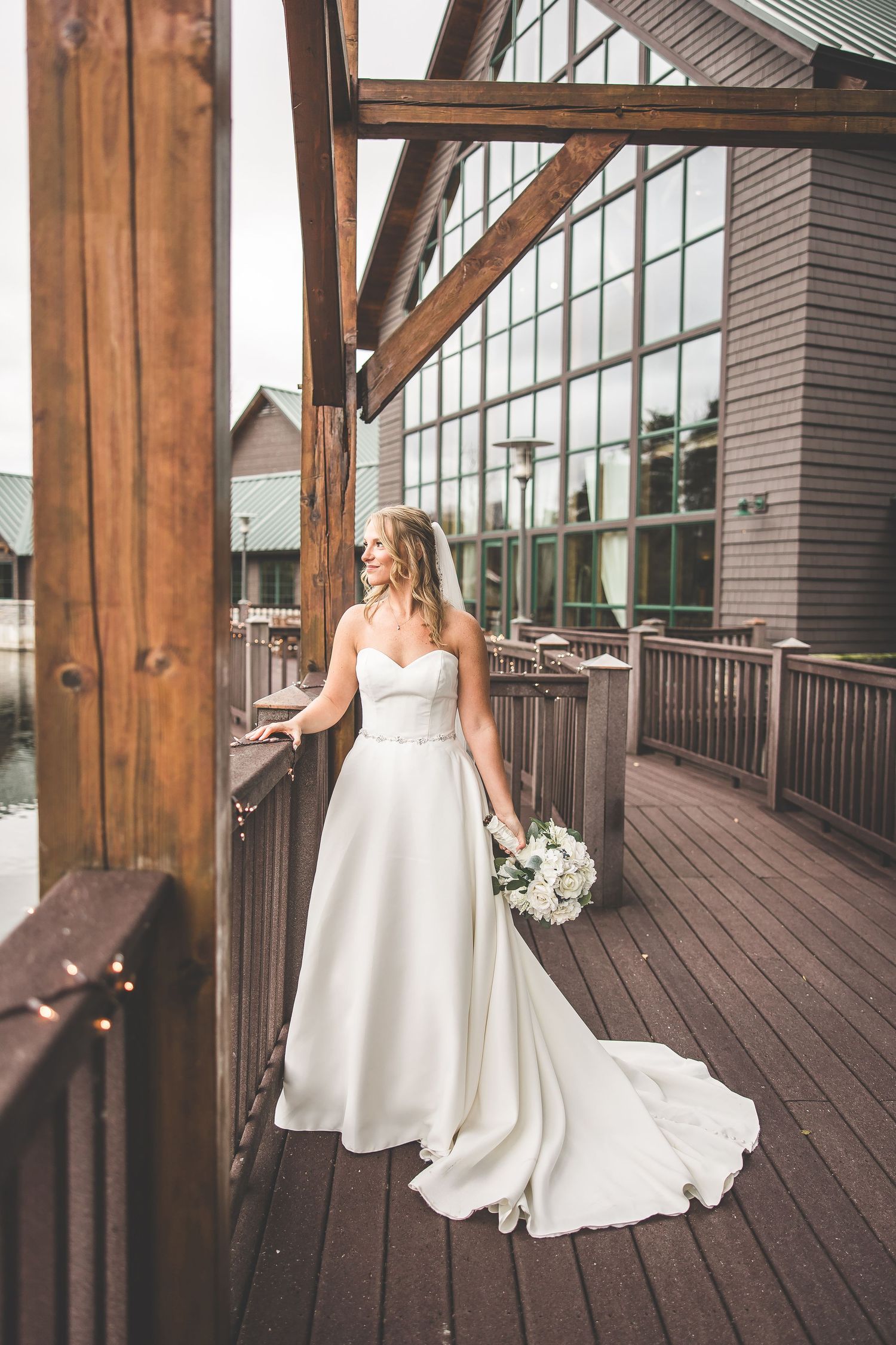 Full-length shot of bride in classic white gown on wooden deck with glass windows.