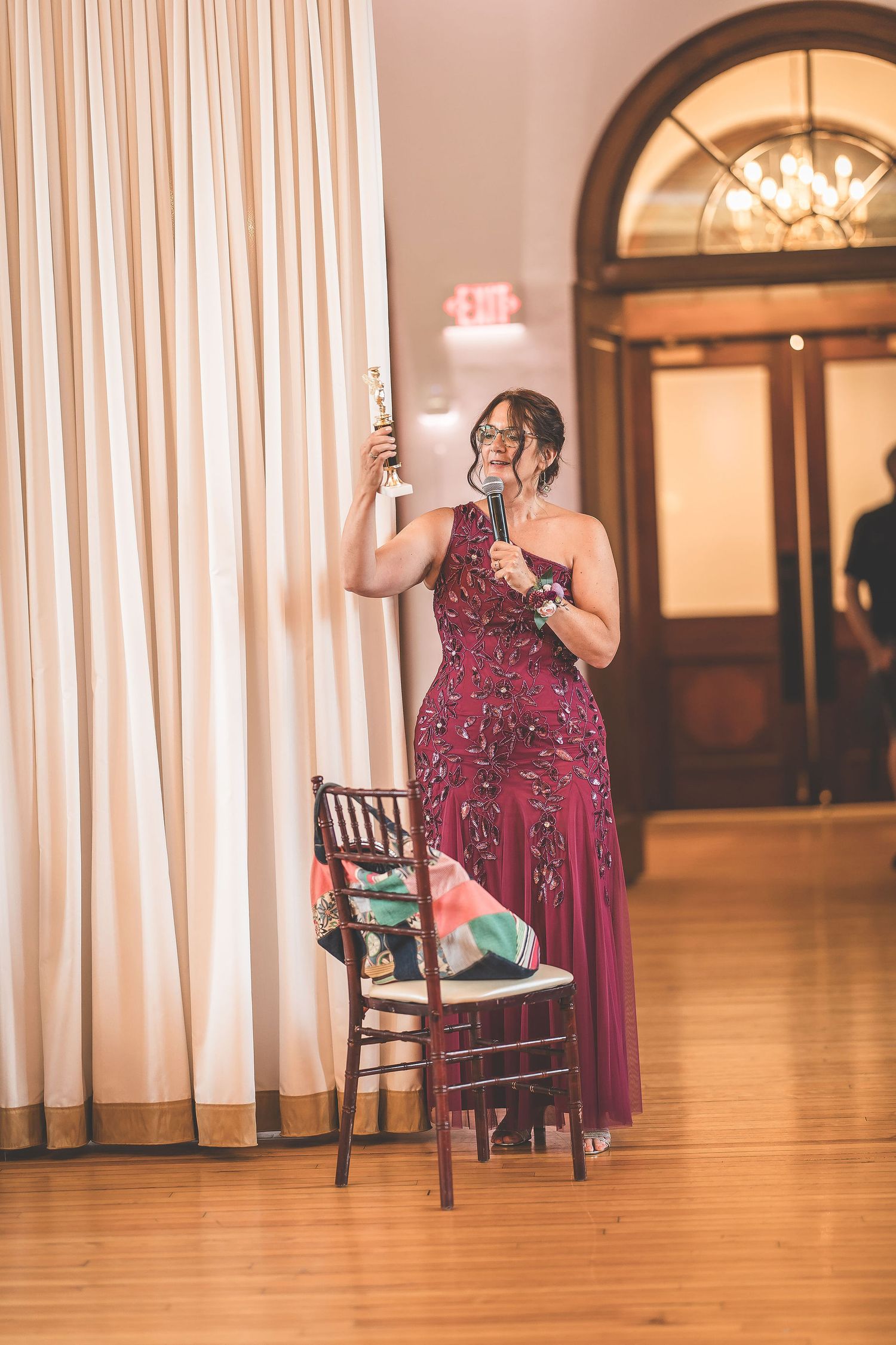 Woman in burgundy sequined dress stands alone in elegant ballroom with hardwood floors and white curtains.
