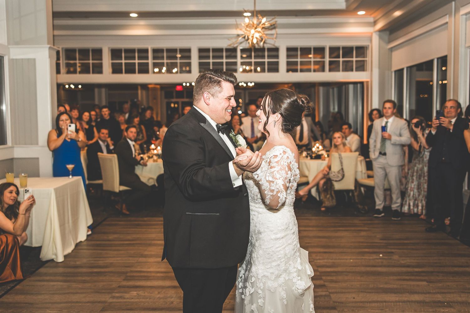 A couple shares their first dance at a wedding reception in an elegant venue with hardwood floors and large windows.