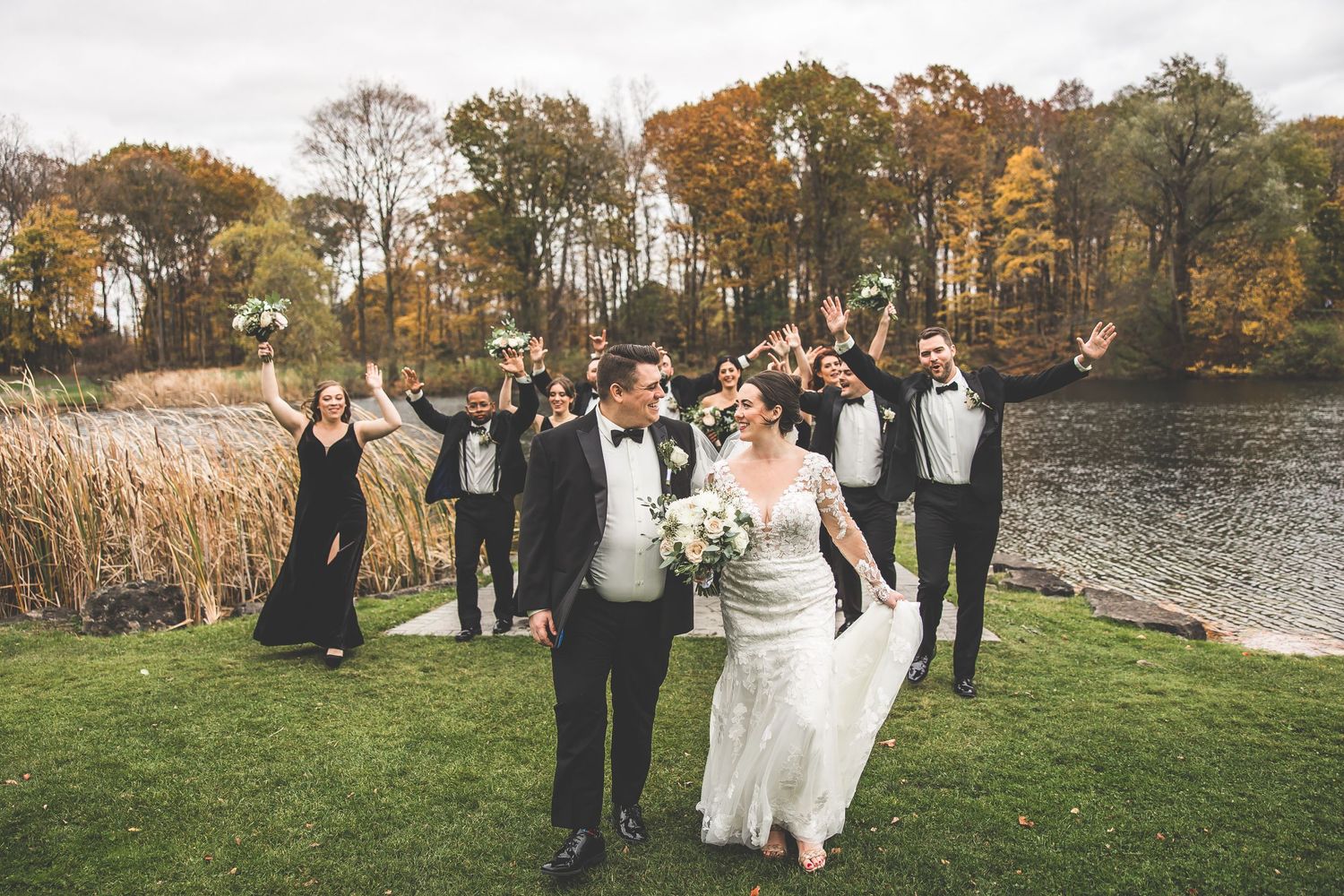 Wedding celebration group walking across grassy lawn near waterfront in fall setting.