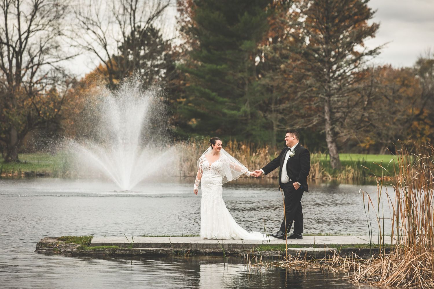 Wedding couple poses by lake with fountain and fall foliage in background.