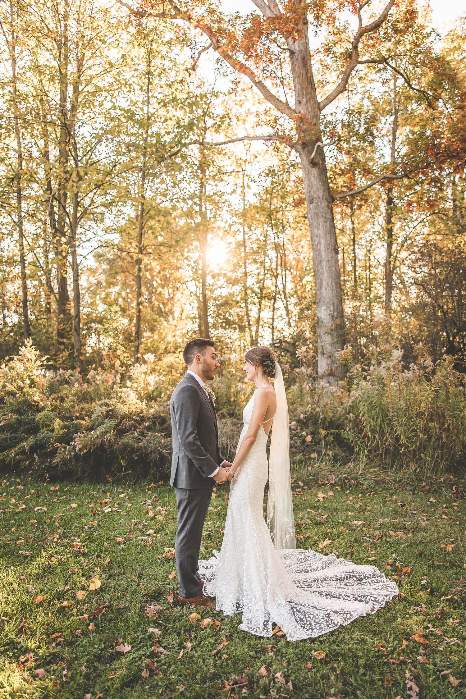 Elegant lace wedding dress train spreads across fallen leaves in autumn woodland setting.