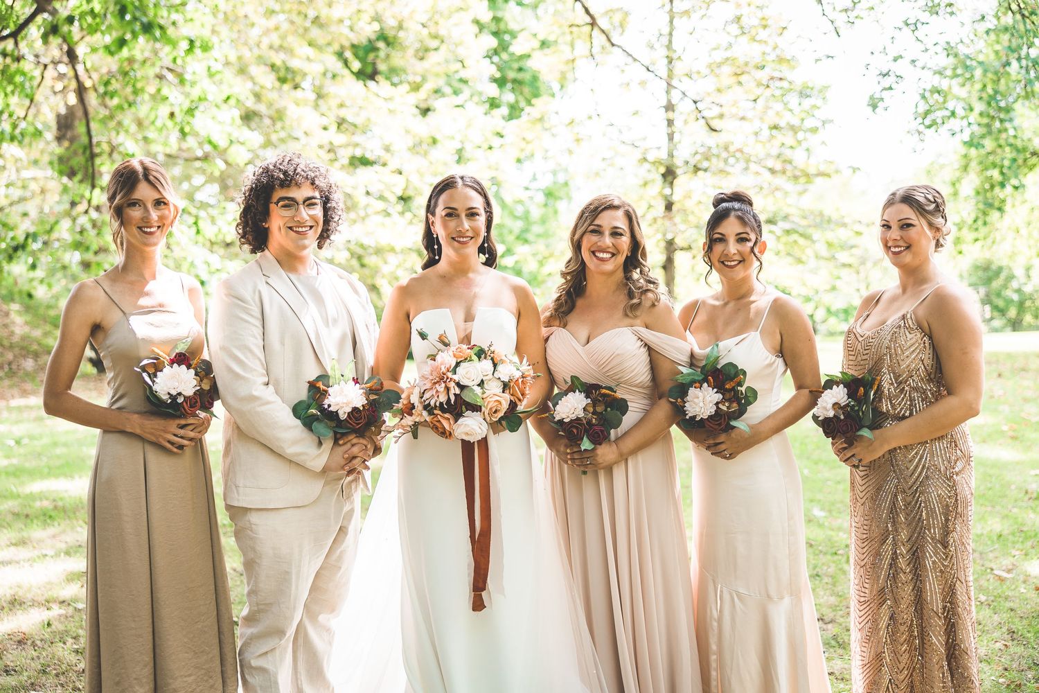 Wedding party portrait outdoors with bridesmaids in neutral dresses holding floral bouquets.