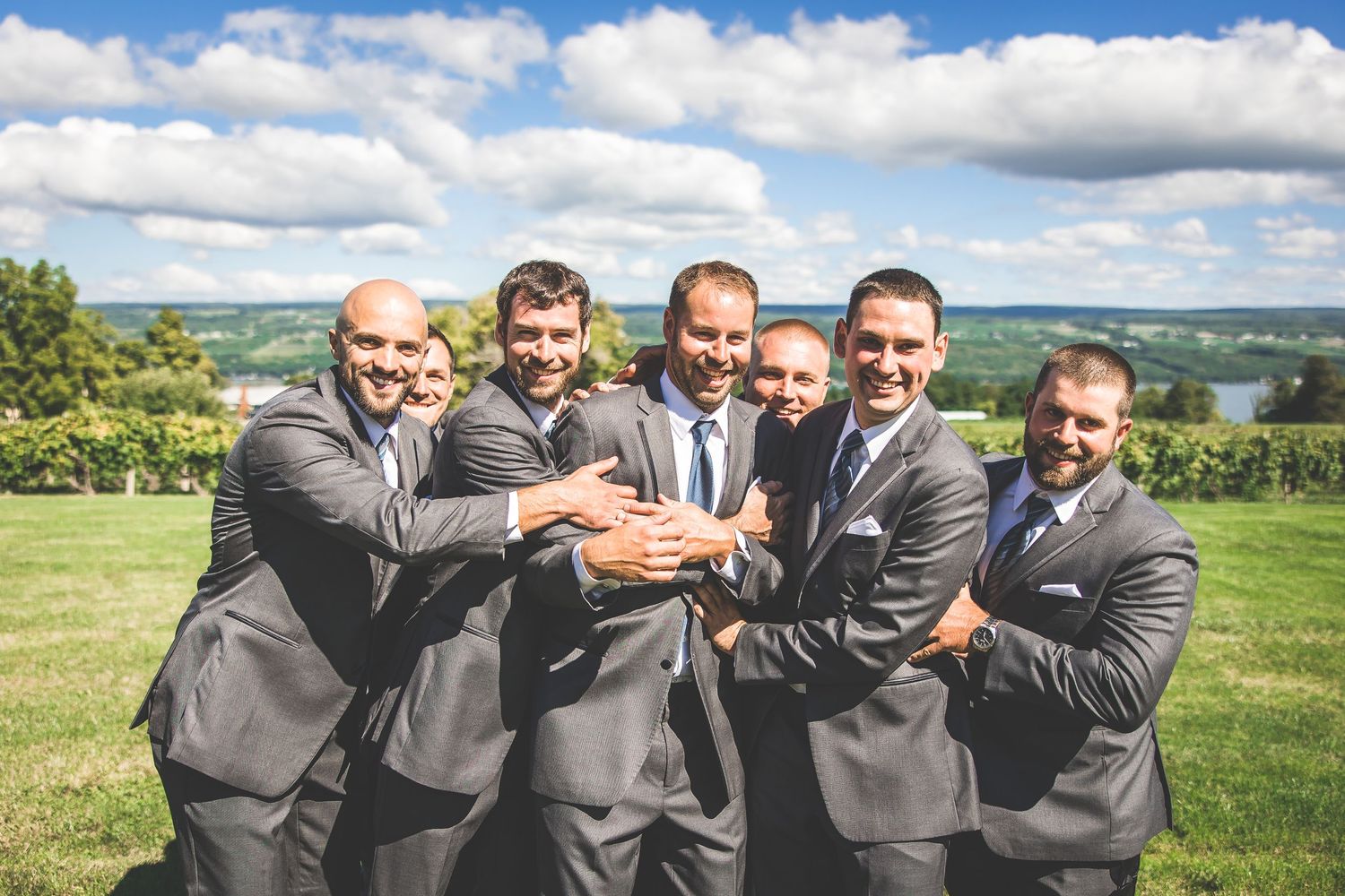 Wedding party in formal attire posing playfully on a grassy hillside with scenic valley view.