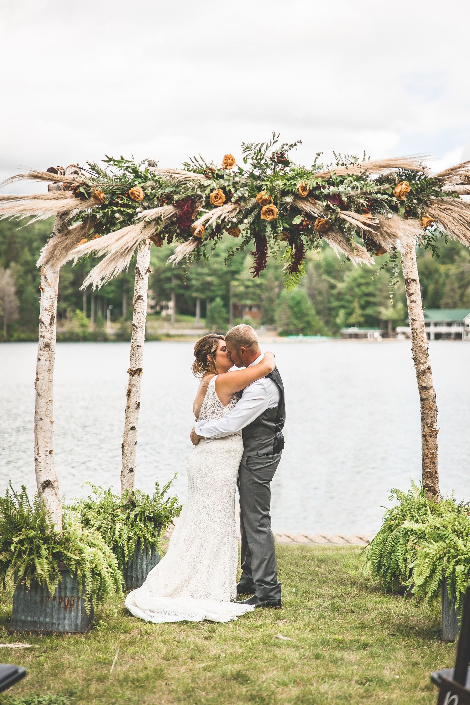 Romantic embrace under floral-decorated birch arch with lake and mountains in background.