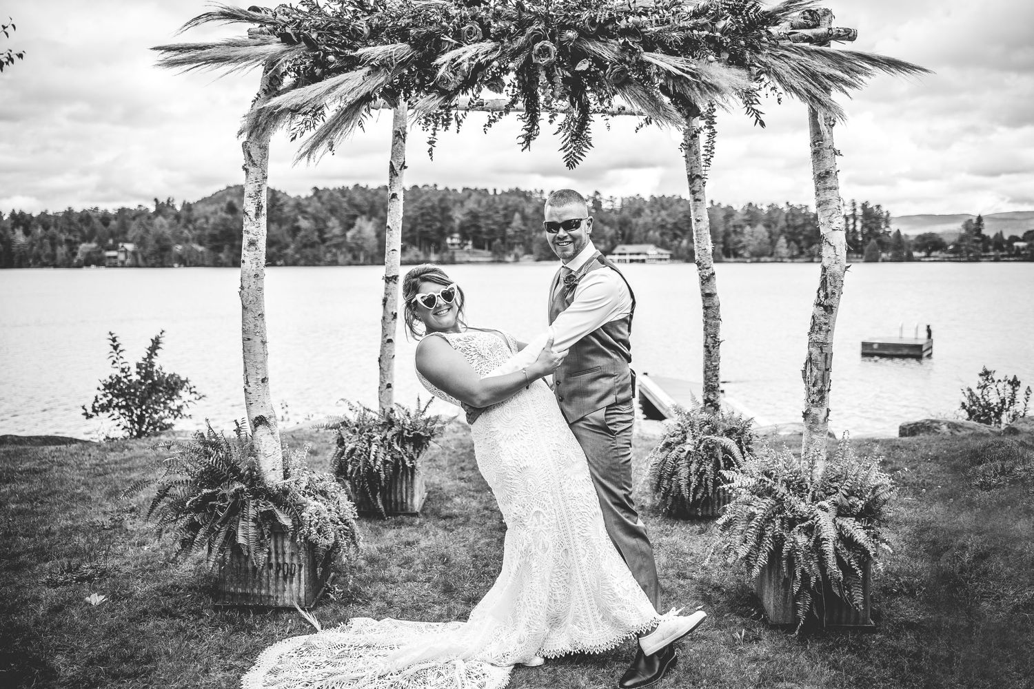 Black and white photo of lakeside wedding ceremony with rustic birch branch arch.