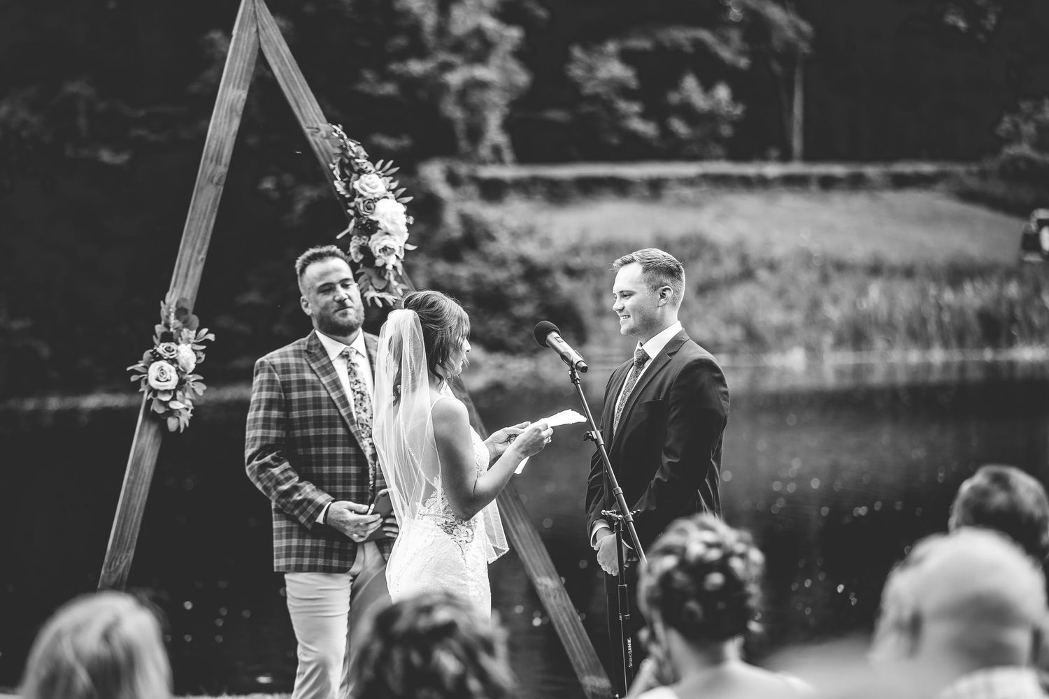 Black and white photo of wedding ceremony by lake with wooden triangle arch.