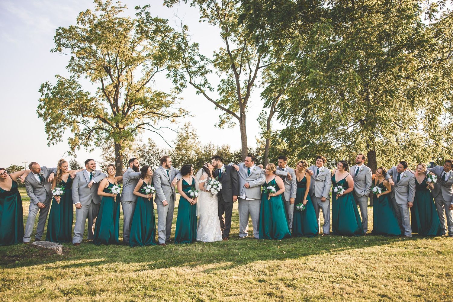 Large wedding party wearing teal dresses poses together on a hillside at sunset.