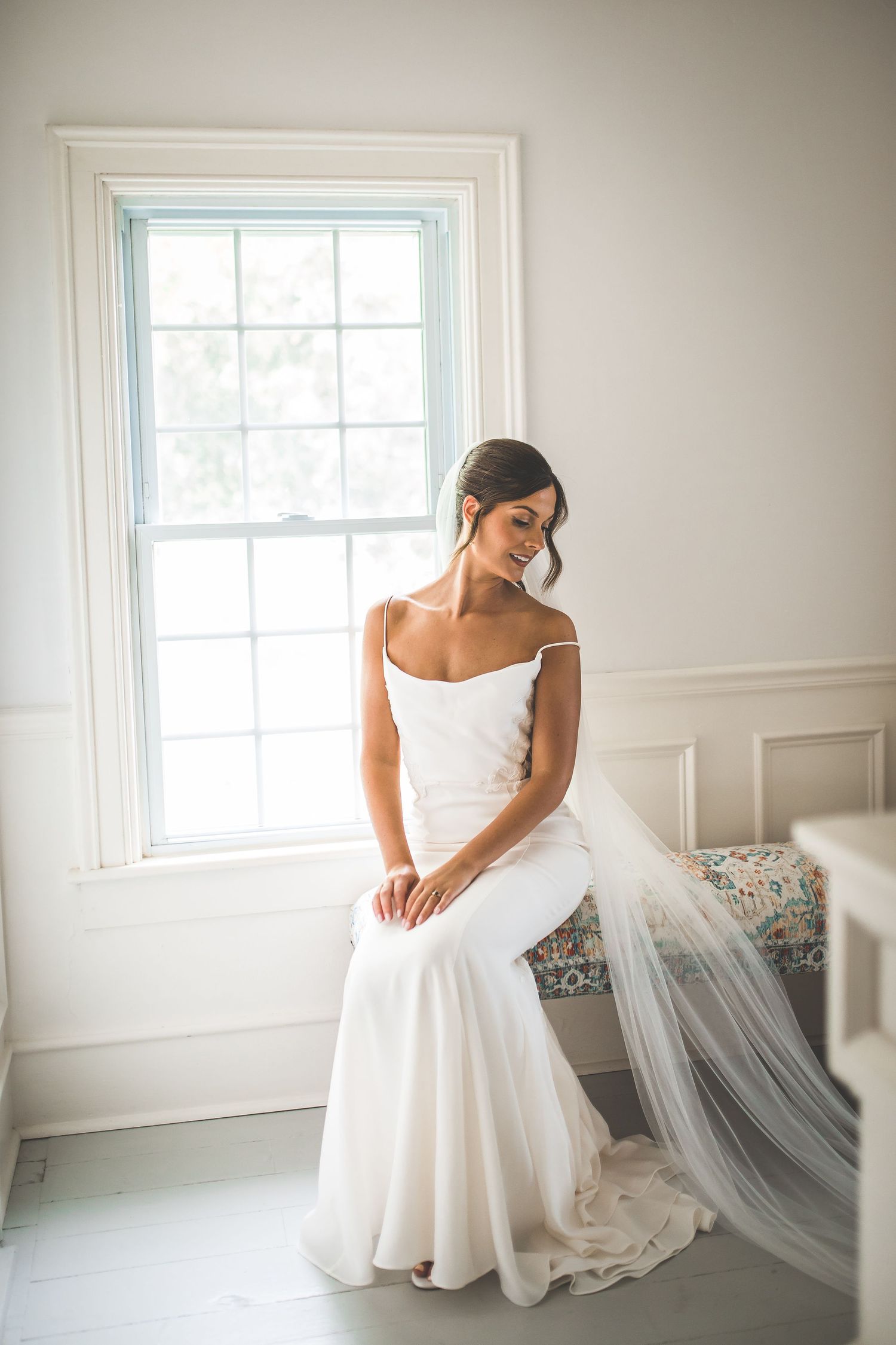 Bride in white dress sits gracefully by window in bright room with natural light.