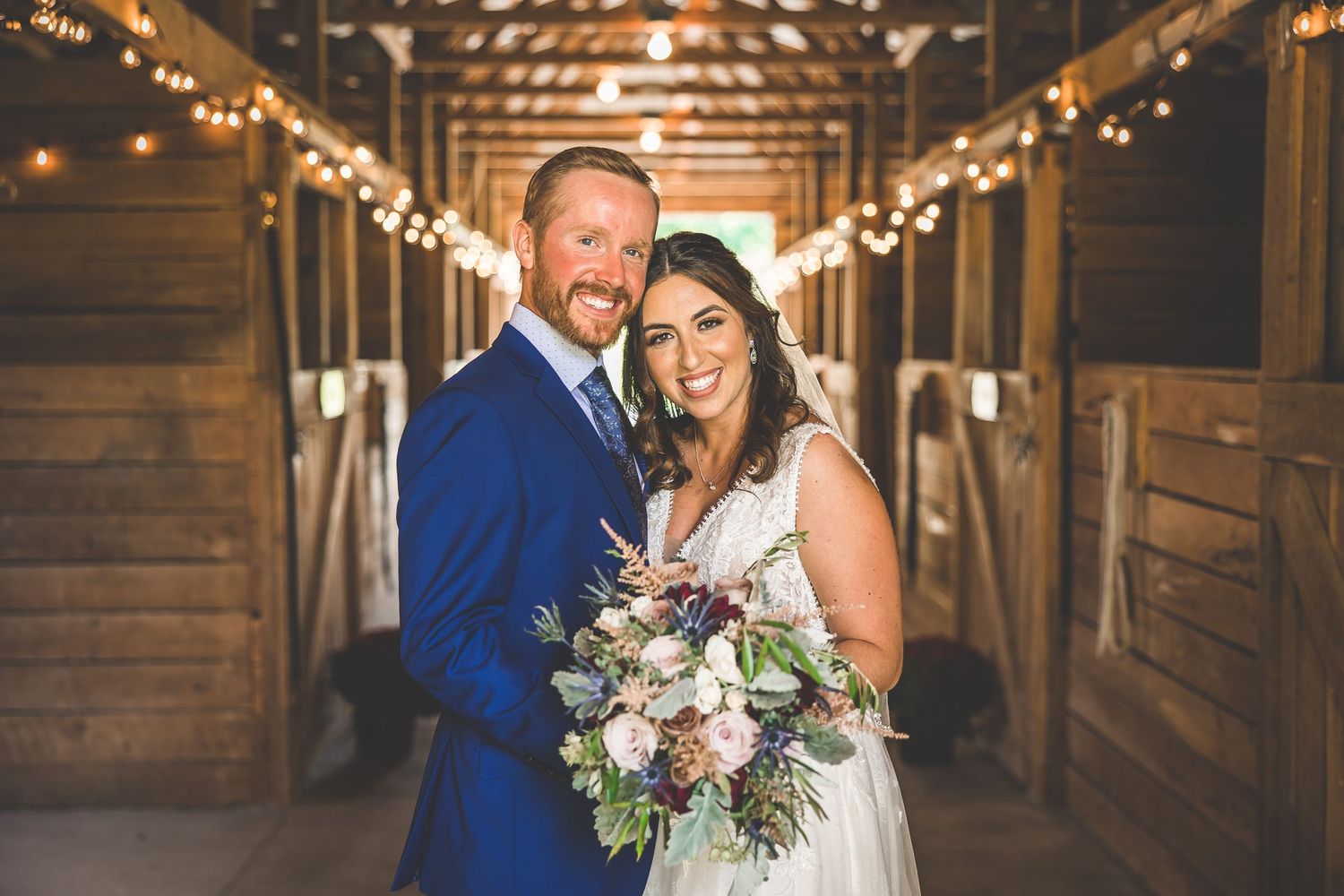 Wedding couple poses in rustic barn venue with string lights and wooden details.