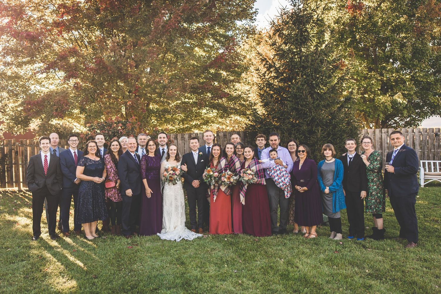 Large wedding group photo with guests in formal attire against fall foliage backdrop.
