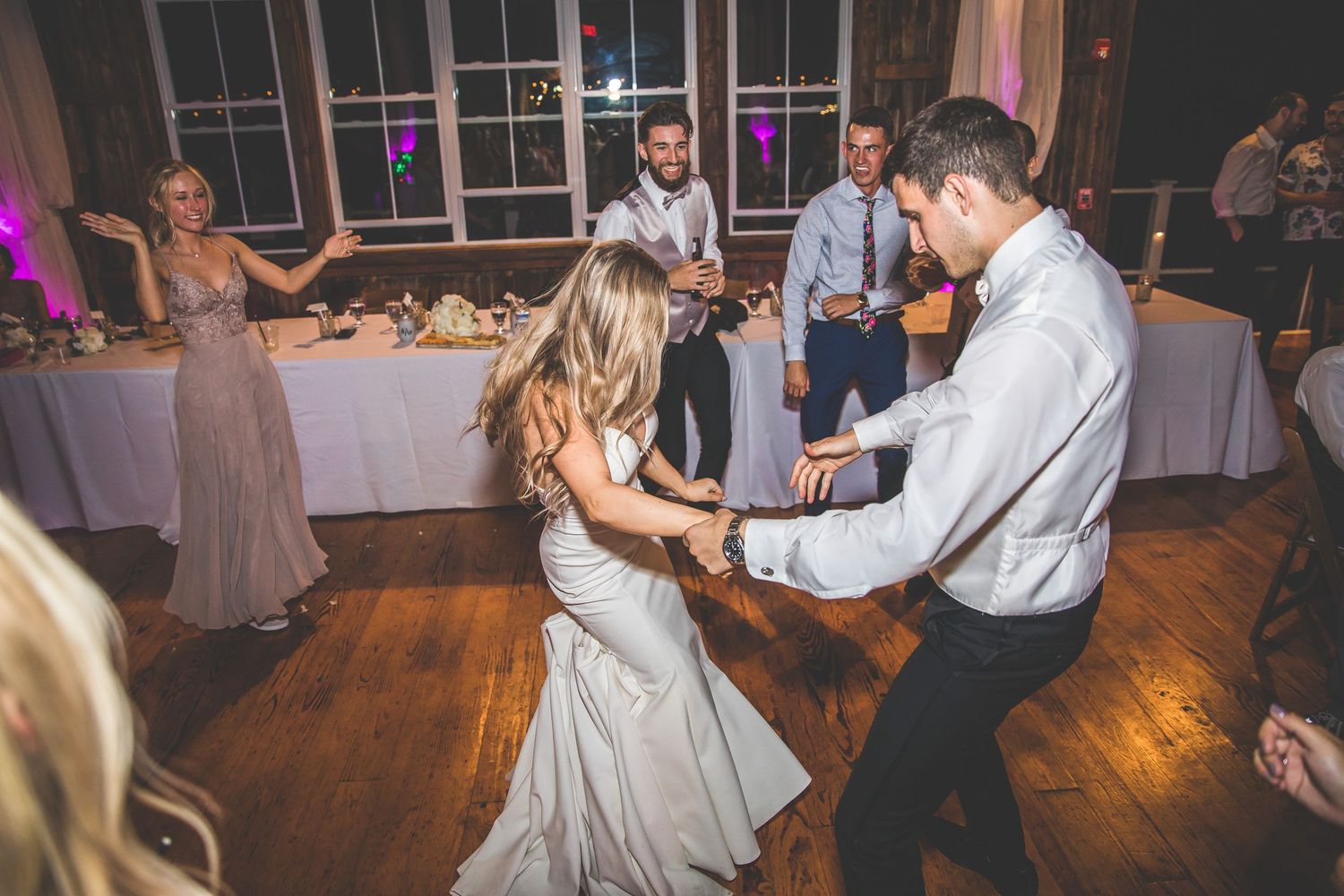 Wedding guests dance and celebrate on a wooden dance floor at an indoor reception venue.