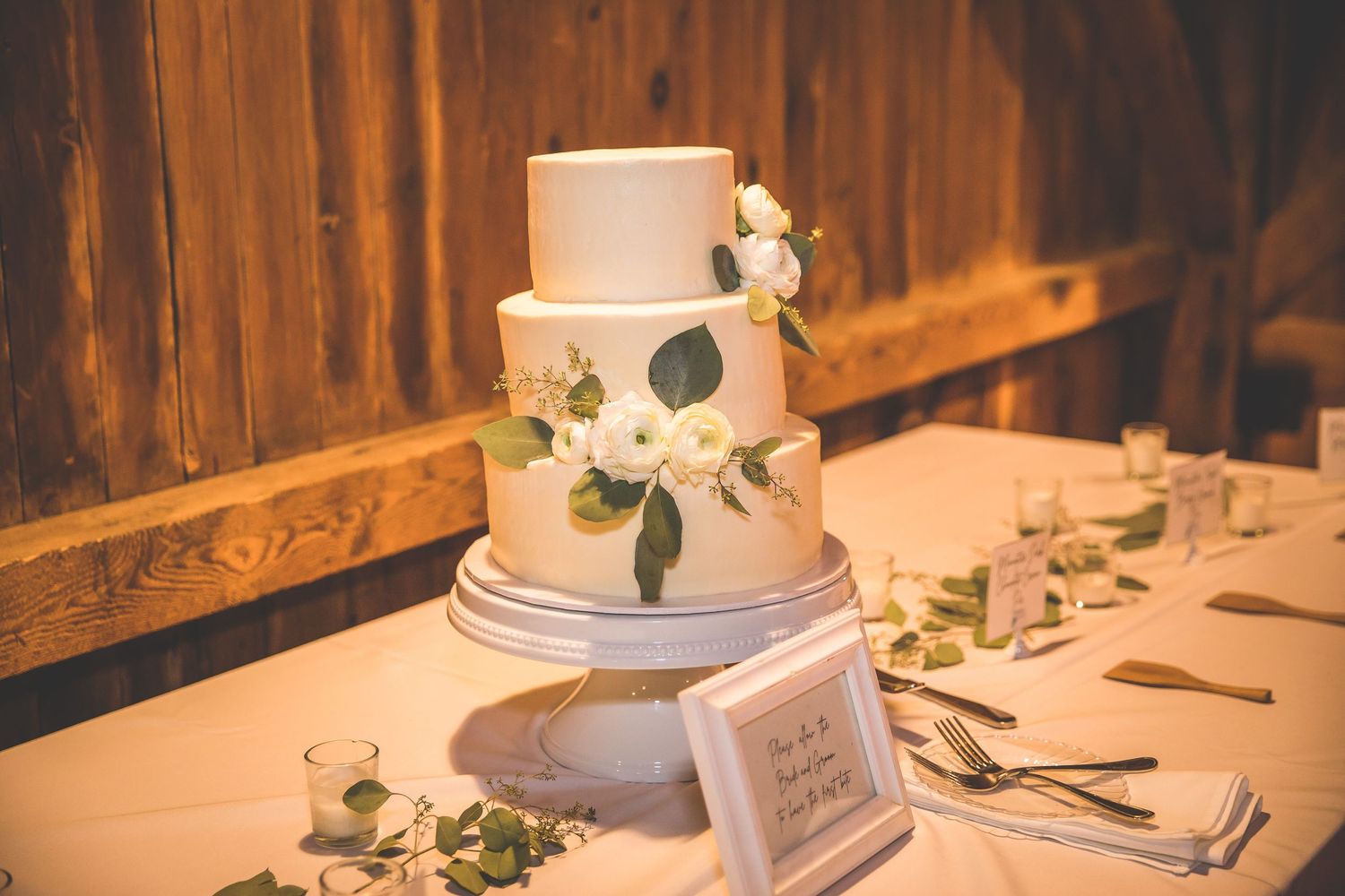 Elegant wedding cake display with white frosting and greenery decorations in a rustic barn setting.