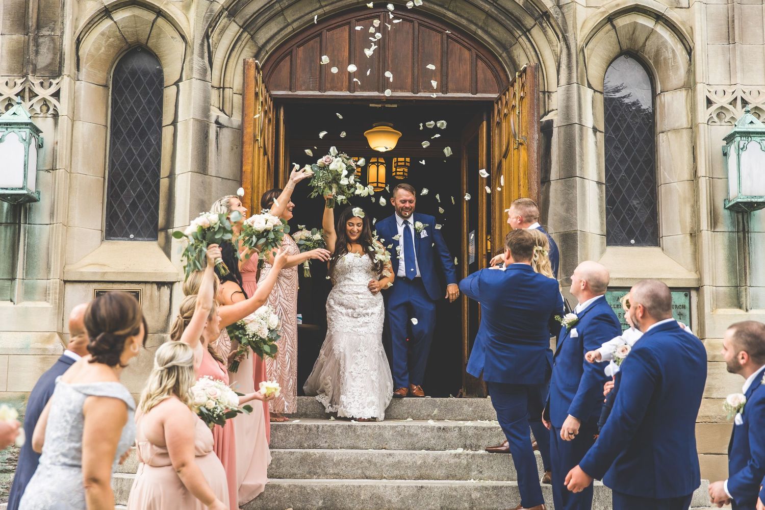 Wedding party exits church as guests throw confetti in celebration outside gothic stone architecture.