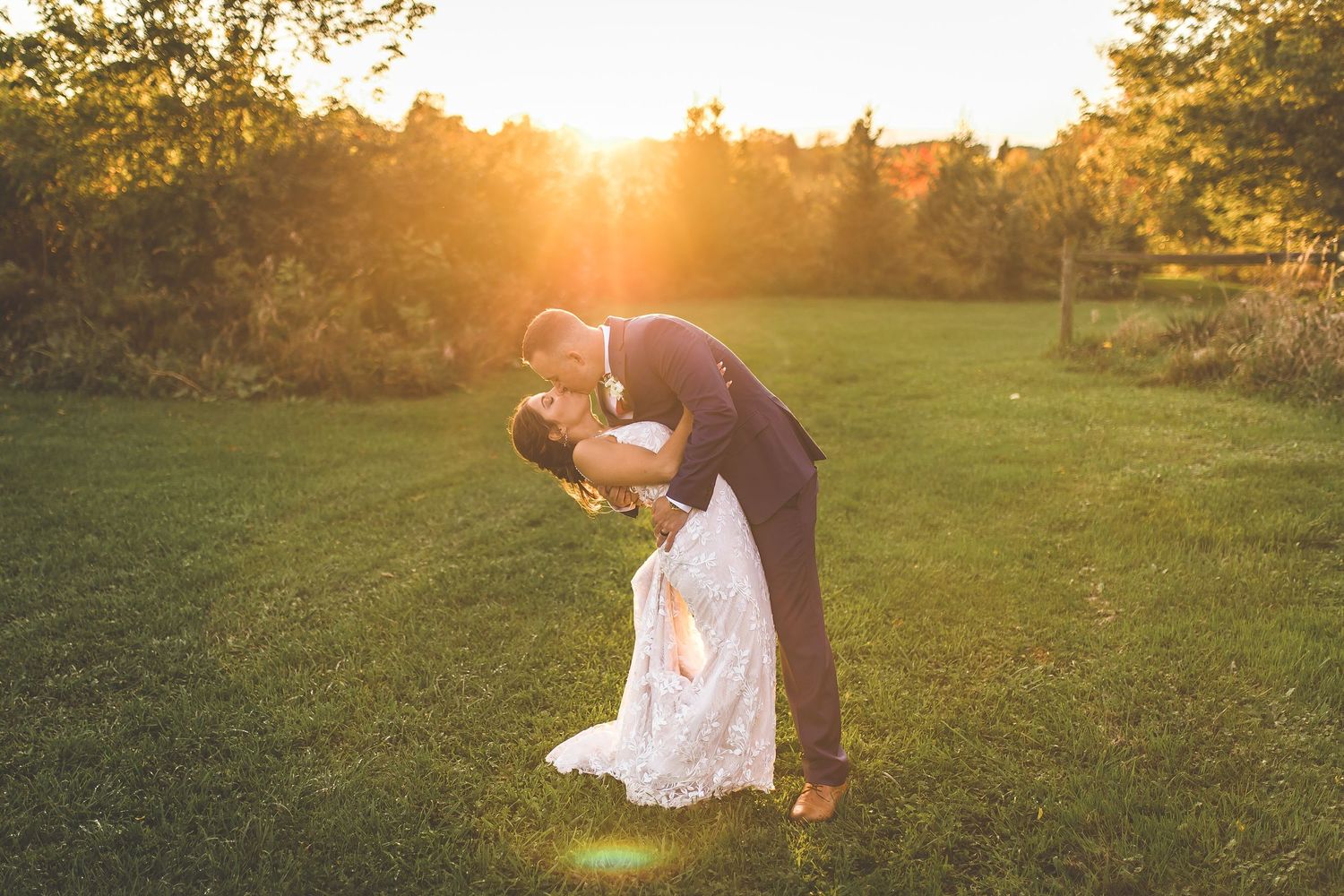 Romantic sunset dip kiss between couple in formal attire on grassy field.