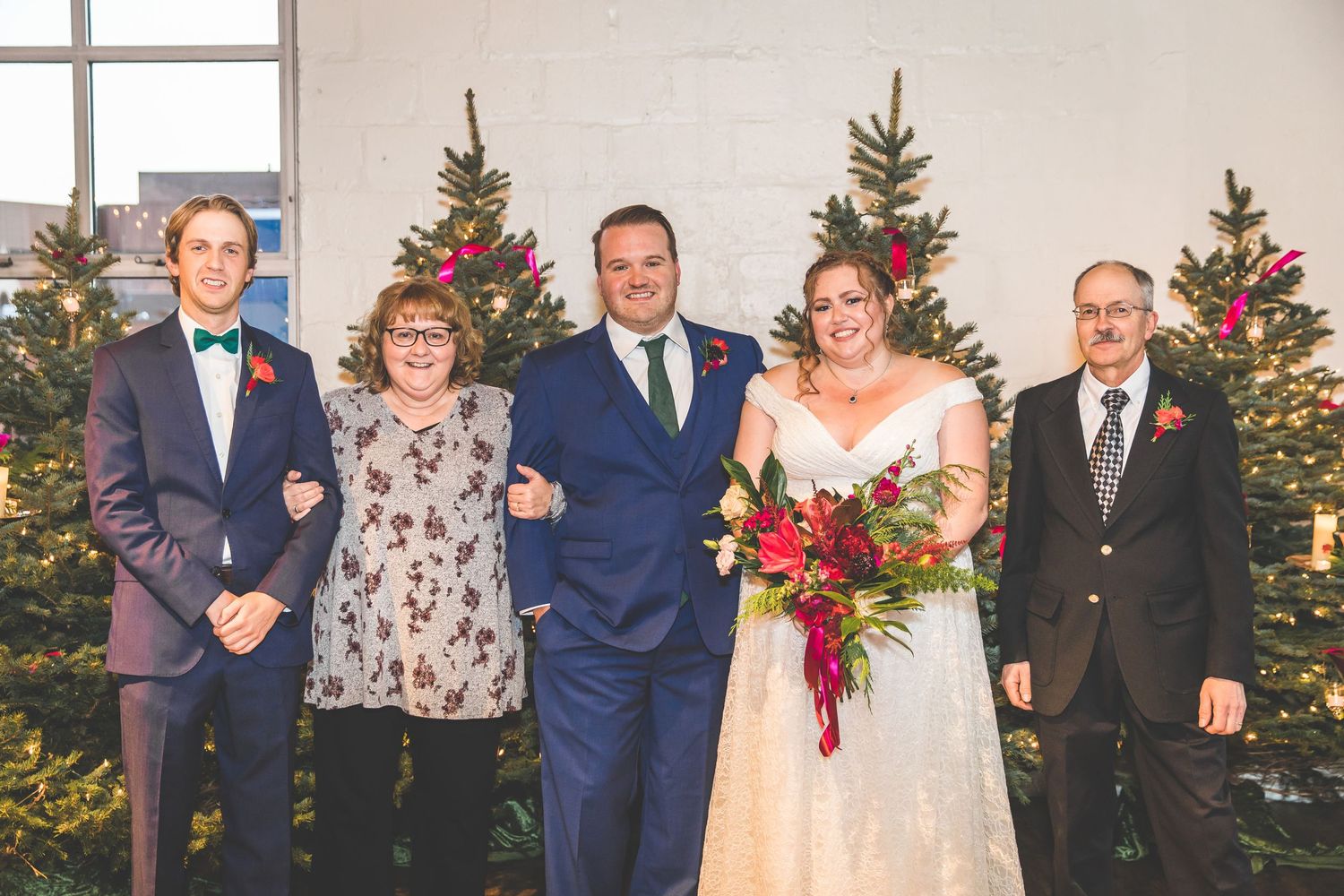 Wedding party members pose formally in front of decorated evergreen trees.