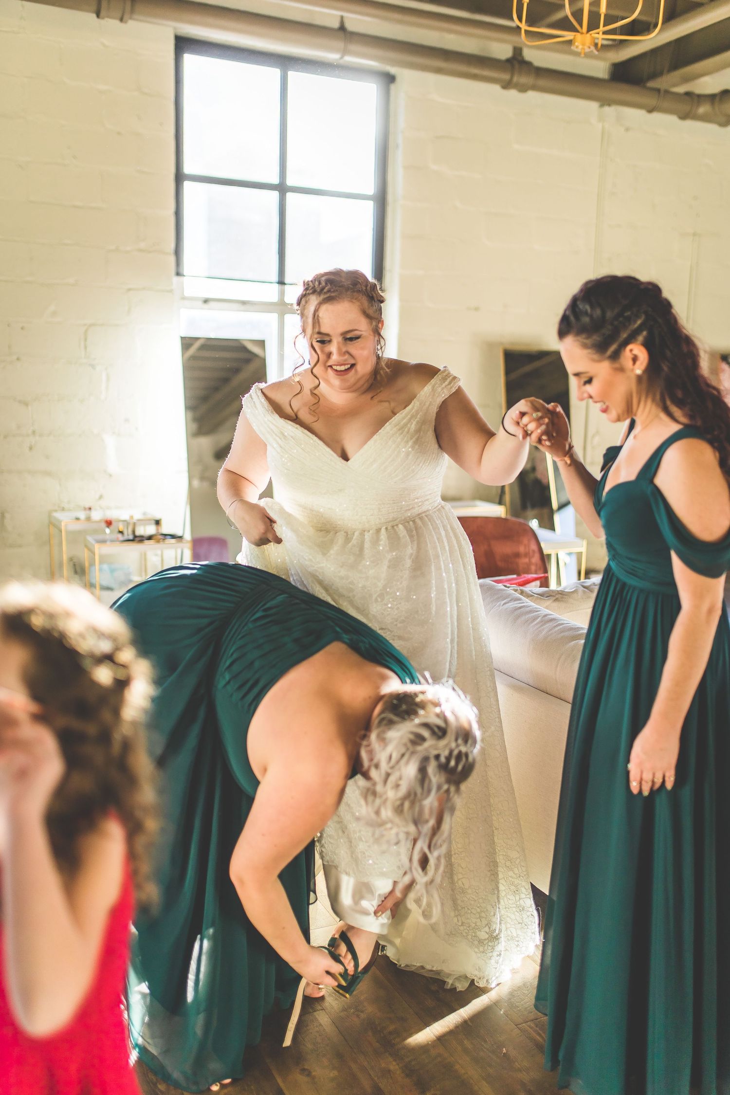 Wedding party getting ready in room with emerald green bridesmaid dresses and white bridal gown.