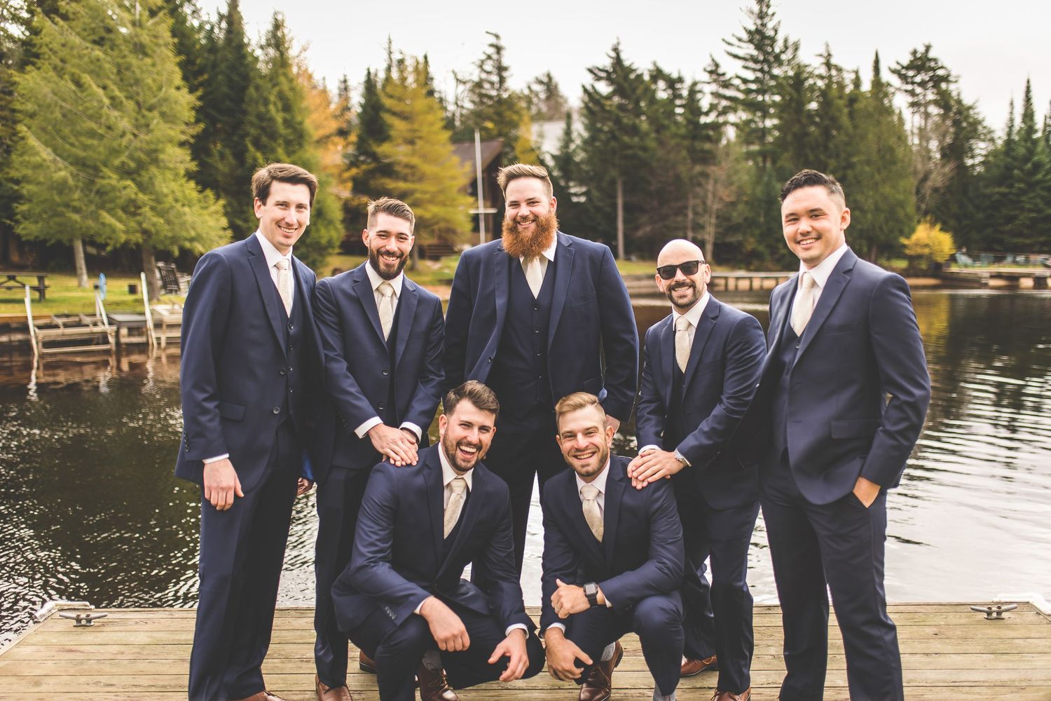 A group in matching navy suits pose together on a wooden dock by a lake surrounded by evergreen trees.