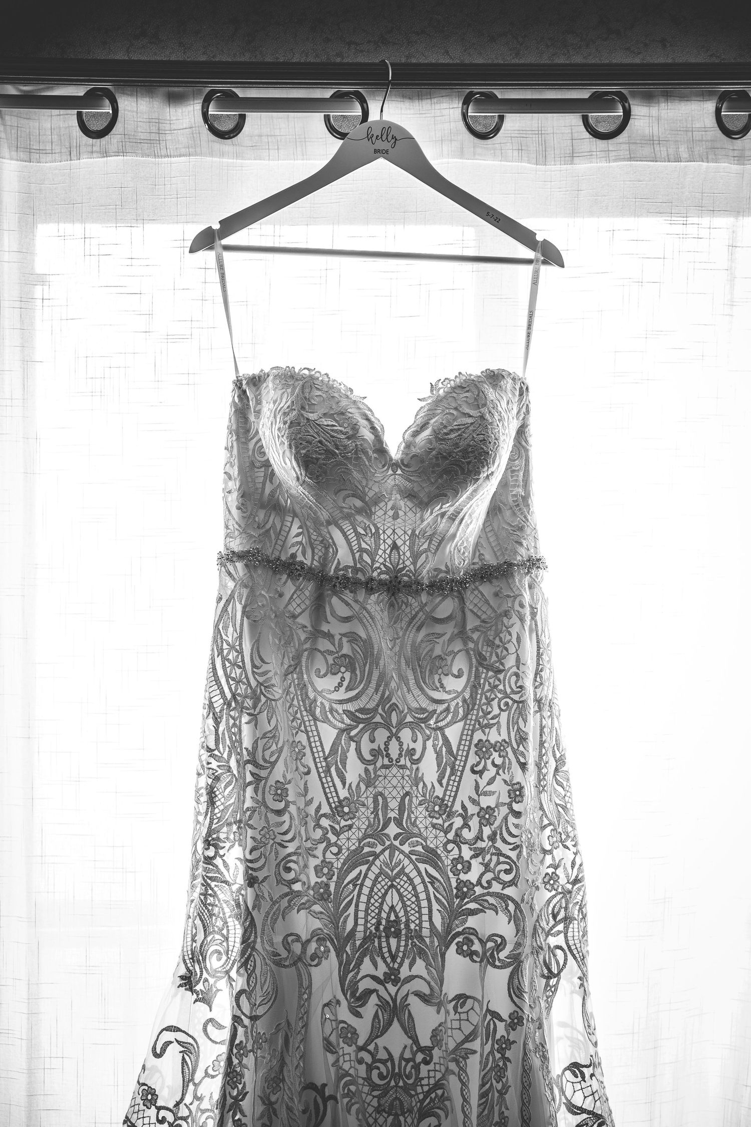 Detailed lace wedding dress hanging against white curtains in window light.