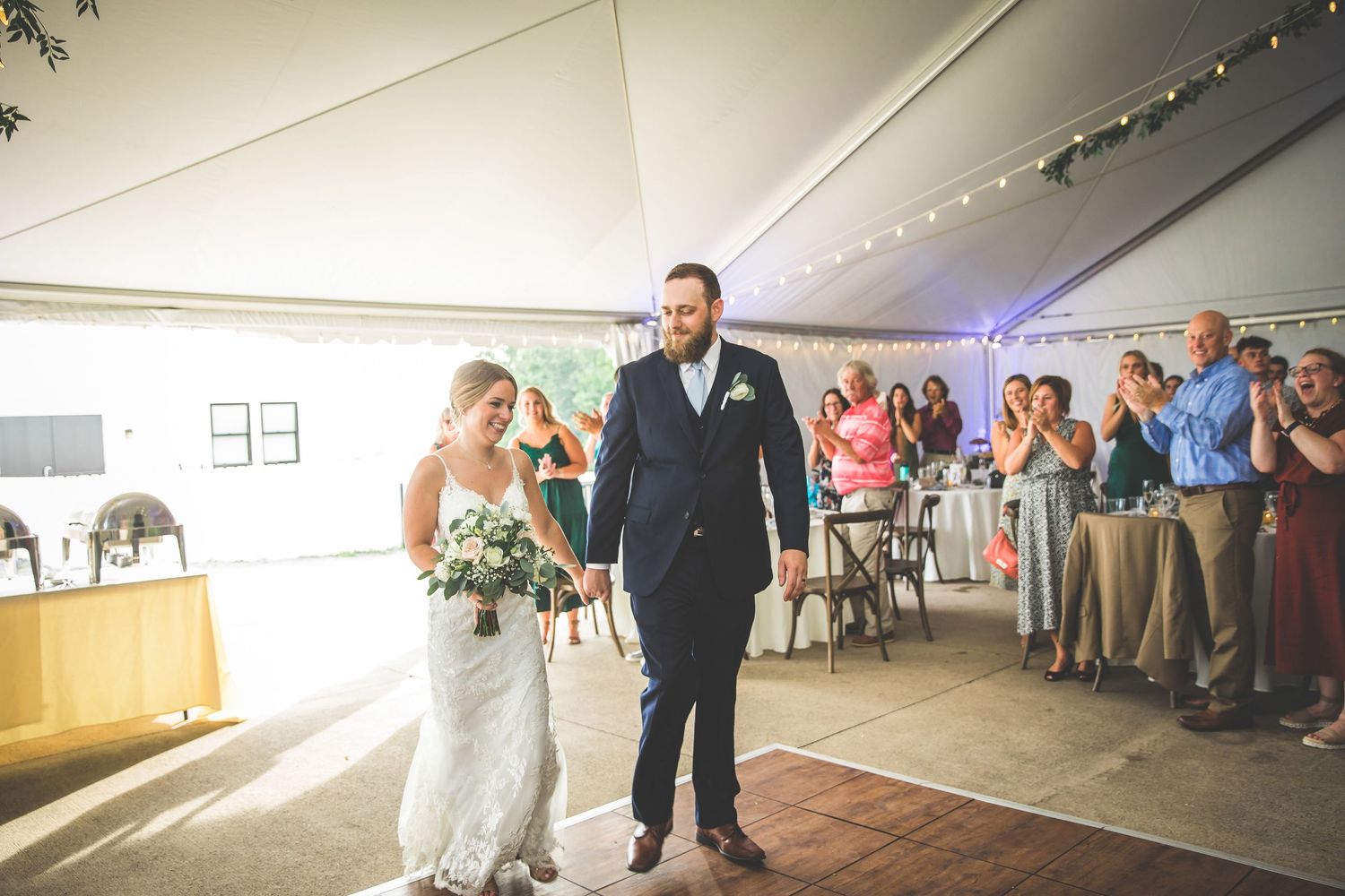 Wedding couple dances together on a rustic wooden floor under a white tent with guests watching.