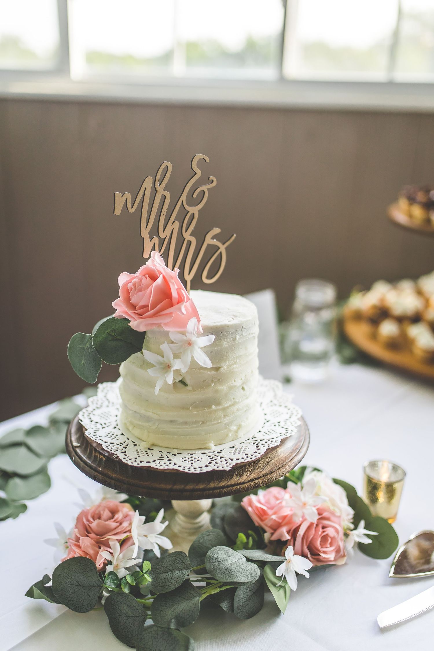 Small white wedding cake decorated with pink roses and a gold Mr & Mrs topper on a wooden stand.