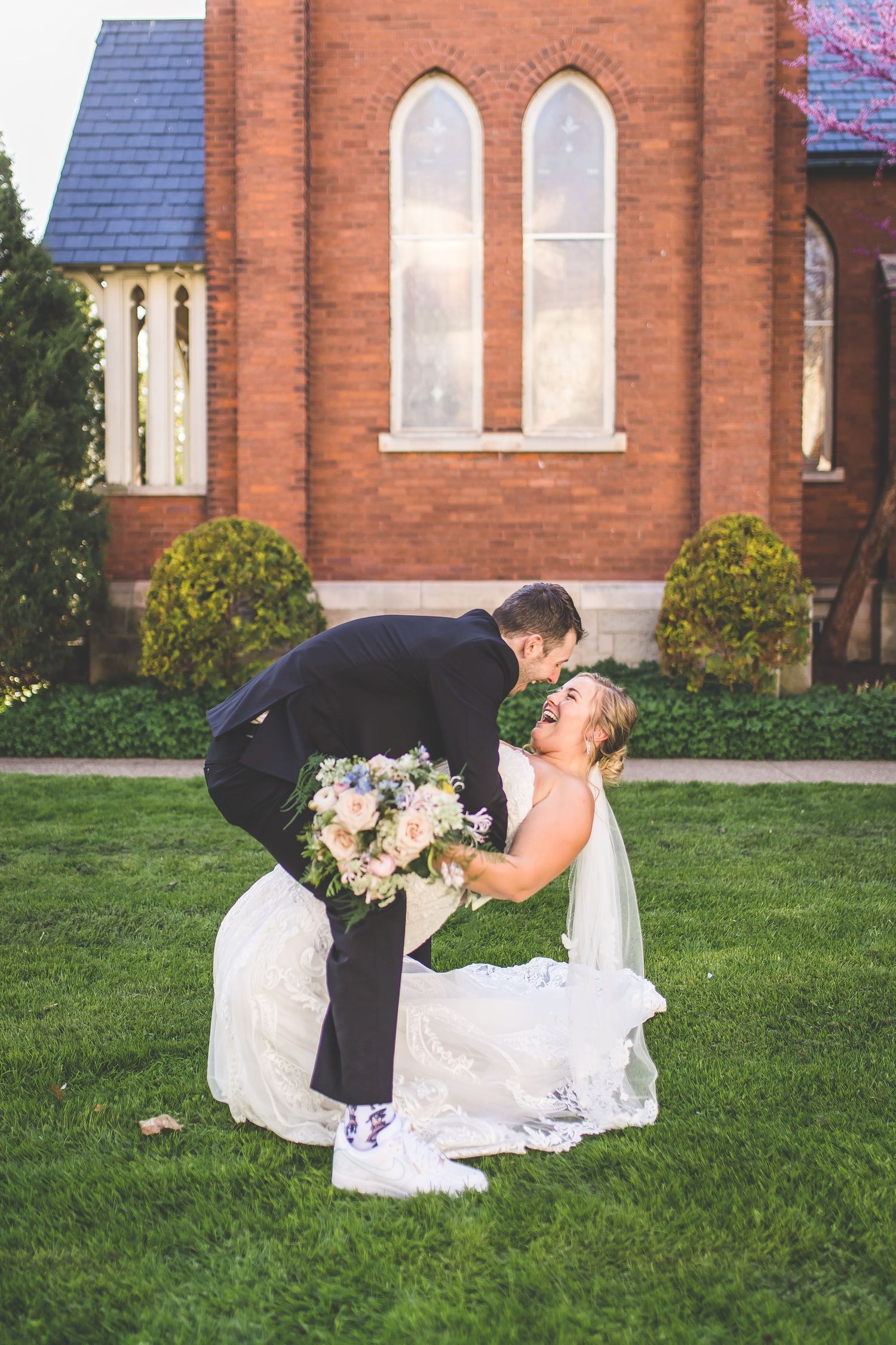 Romantic wedding couple shares playful moment on church lawn with red brick building in background.
