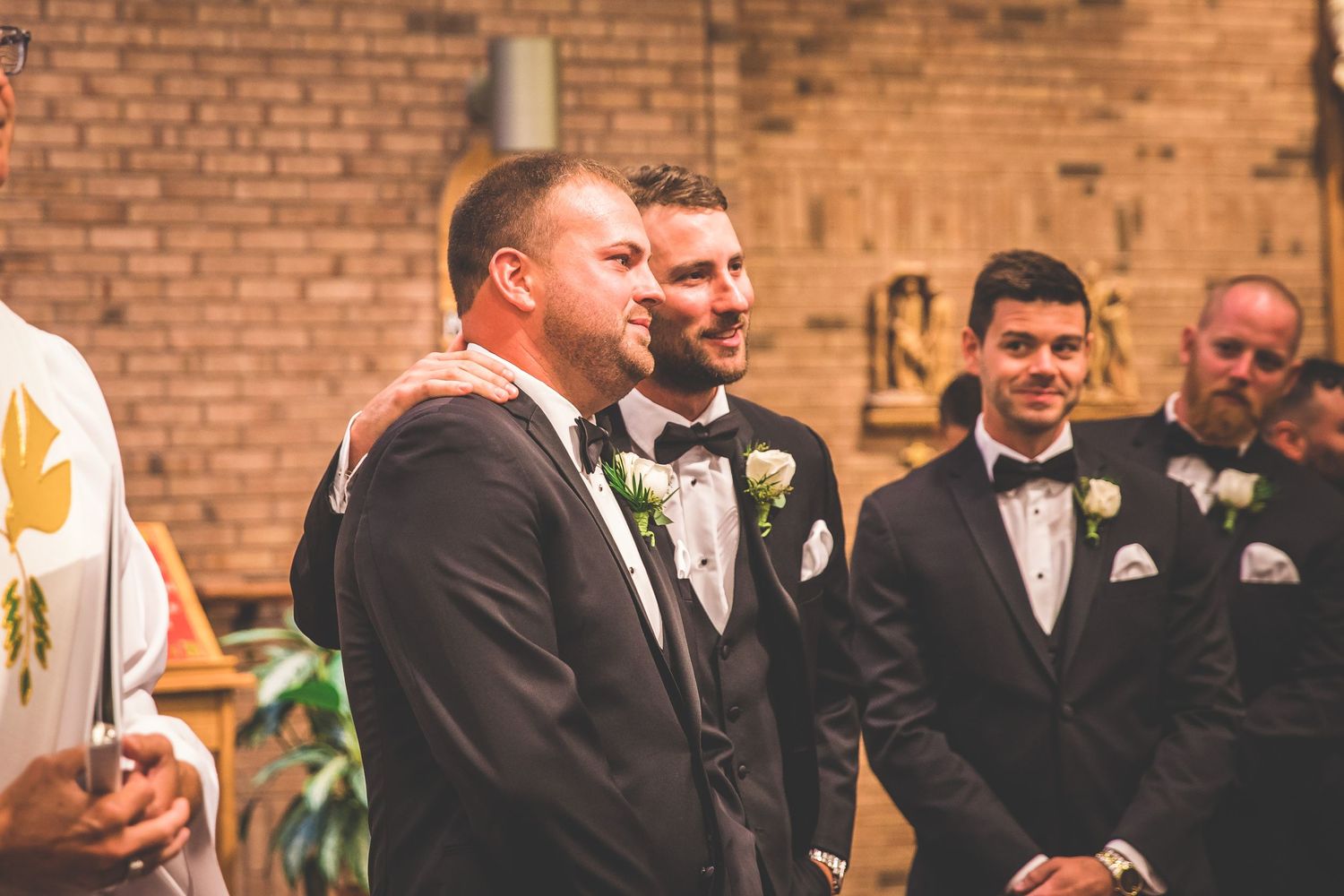 Wedding party members in formal attire gather at a brick-walled venue during a celebration.